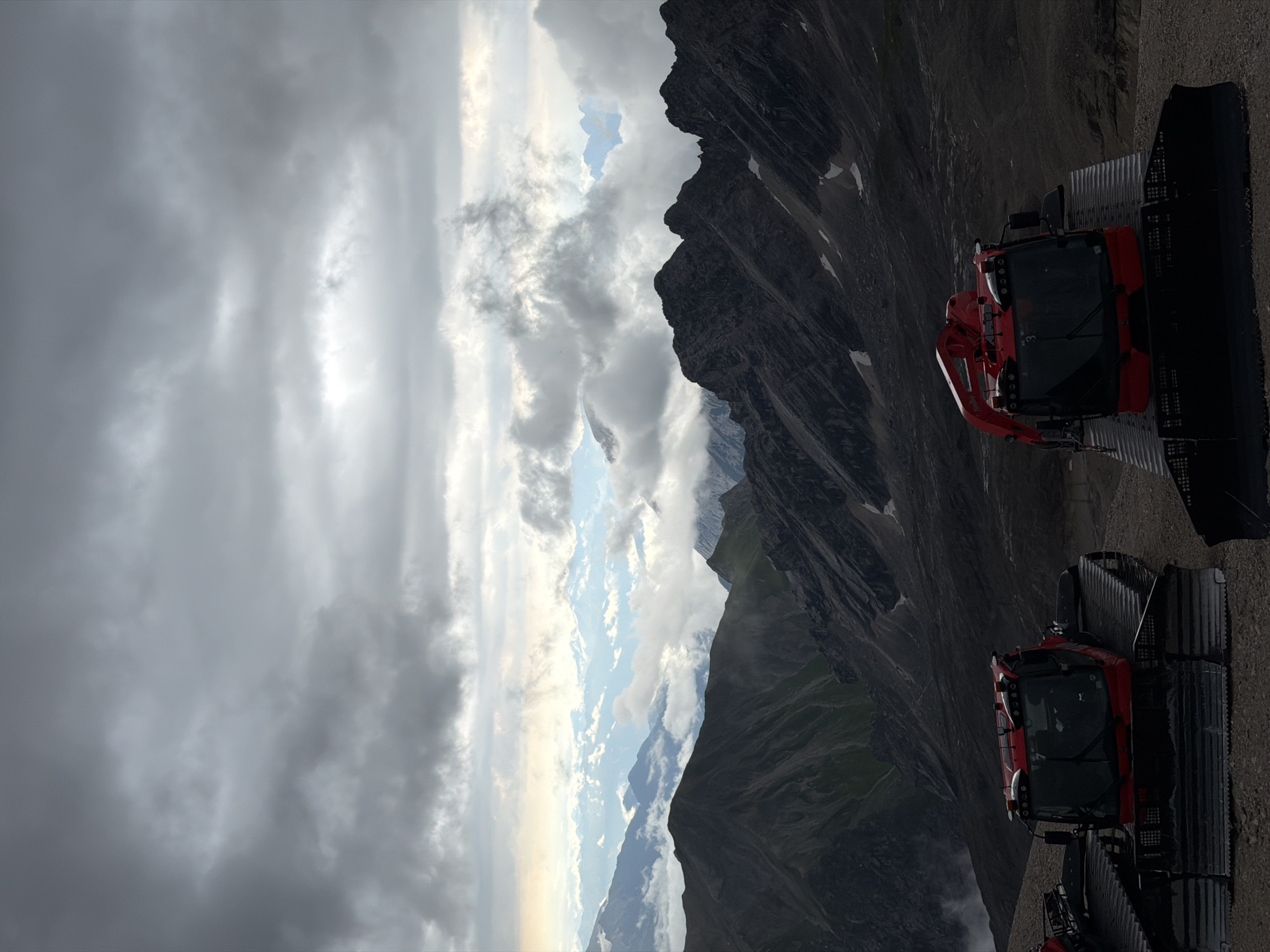 The image depicts a mountainous landscape under a cloudy sky, likely in or near Garmisch-Partenkirchen, Germany, given the presence of snow groomers. In the foreground, two red snow groomers, also known as PistenBully vehicles, are parked on a gravelly surface. The snow groomers have large tracks and prominent headlights. The middle ground features rugged, dark mountain slopes, some with patches of snow, and dramatic rock formations. Beyond these, a vast expanse of layered mountains stretches into the distance, partially obscured by white clouds and a hazy, blue sky. The overall atmosphere is somewhat dramatic due to the overcast conditions and the imposing mountain scenery. There are no people visible in the image.