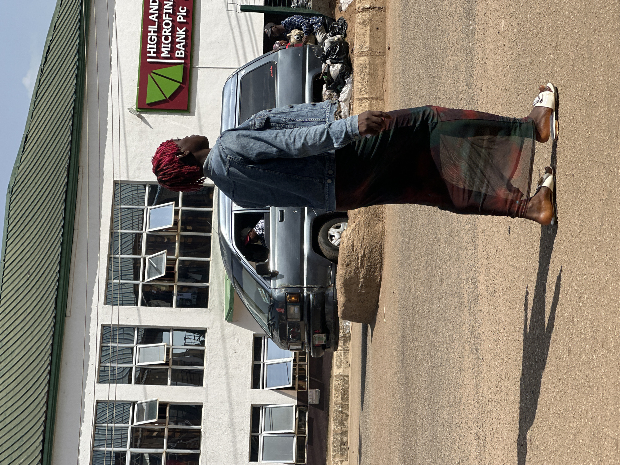 In Bukuru, Nigeria, a person with vibrant red dreadlocks is walking on a dusty asphalt road. They are wearing a denim jacket and a long, sheer, patterned skirt, with white sandals on their feet. In the background, a grey minivan is parked near a building with a green roof and numerous windows. A sign on the building reads "HIGHLAND MICROFINANCE BANK Plc". To the right of the minivan, several large black trash bags are piled on the ground, and two individuals are seated nearby. The scene is illuminated by bright sunlight, casting a distinct shadow of the walking person on the road.