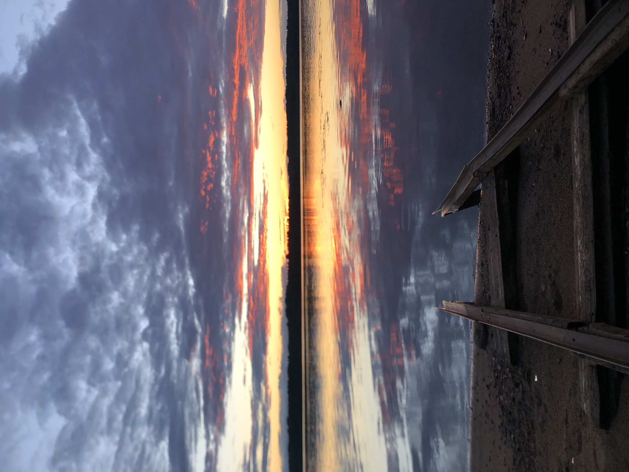 An outdoor, eye-level, vertical, full shot depicts a lake at dusk in Svirstroy, Russia. The sky is filled with dramatic clouds that are a mix of dark grey and vibrant pink and orange hues, indicating a sunset or sunrise. The colors from the sky are beautifully reflected in the calm water of the lake, creating a mirror image. In the foreground, there is a sandy shore with a wooden ramp or track structure leading towards the water. This structure appears to be made of weathered wood and metal, possibly for launching or retrieving a boat. The distant shoreline is lined with a dark silhouette of trees. There are no people visible in the frame.