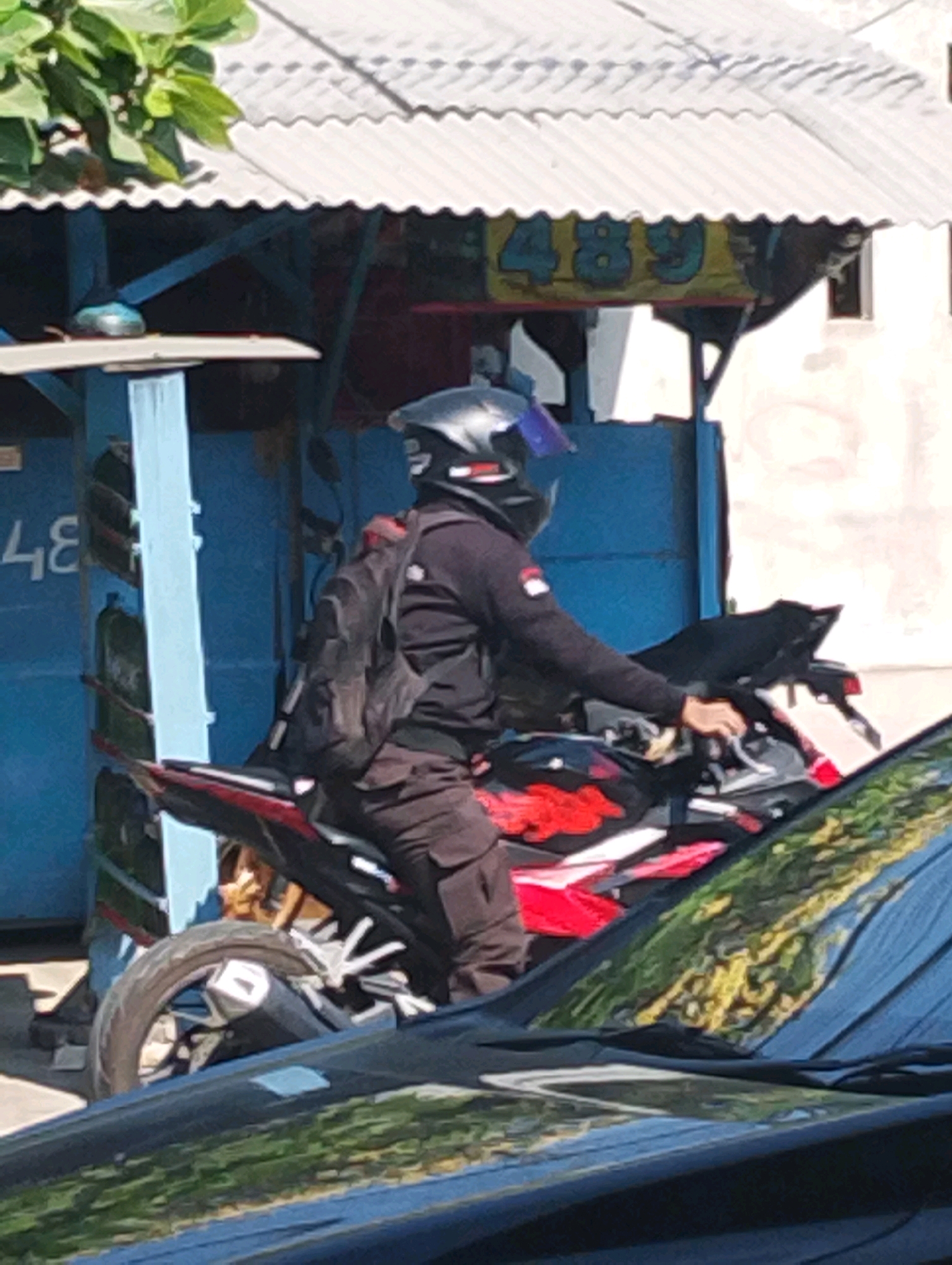 In Manaruwi, Indonesia, a person wearing a black helmet and a black jacket with a backpack is riding a red and black motorcycle. The rider is on a street with a blue building in the background. A sign with the numbers "489" is visible on the building. The motorcycle has a red and white design on its side. The scene is captured from a low angle, with a portion of a dark-colored car visible in the foreground. The environment appears to be a sunny day.