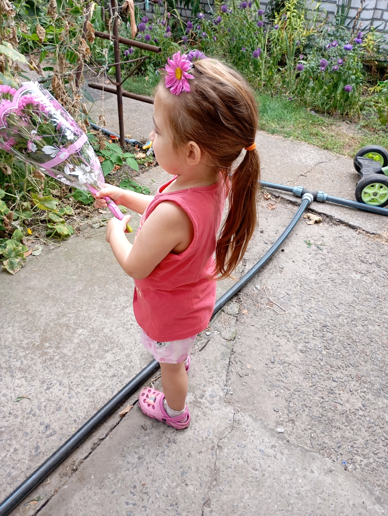 A young girl, likely between 3 and 4 years old, stands outdoors on a concrete path, holding a bouquet of pink flowers wrapped in clear plastic. She is wearing a pink sleeveless top and light-colored shorts with a pink pattern, along with pink slip-on shoes. A pink flower is clipped into her hair, which is pulled back into a ponytail. The background features green foliage, purple flowers, and a brick wall. A black hose and a toy lawnmower with green wheels are also visible on the path. The overall scene suggests a casual outdoor setting, possibly a backyard or garden. The location is identified as Horishni Plavni, Ukraine.