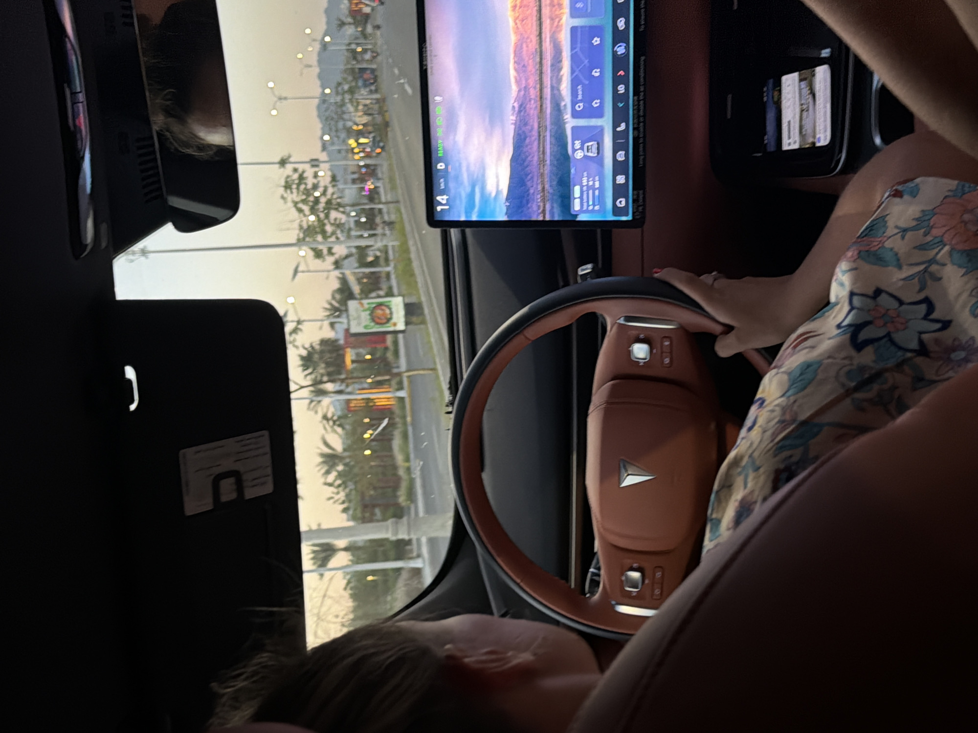 In the city of Sharm El Sheikh, Egypt, a woman is seated in the driver's seat of a modern car, her hands resting on the tan leather steering wheel. The car's interior is visible, featuring a large, central touchscreen display that shows a scenic landscape of mountains and a body of water, along with navigation information. The screen displays the current speed as 14 km/h and indicates the car is in "READY" mode. Outside the car, the view through the windshield reveals a street scene with palm trees, streetlights, and buildings, suggesting an urban or suburban environment at dusk or dawn. A second person's reflection is visible in the rearview mirror, indicating they are in the car. A phone is charging on a console in the car.