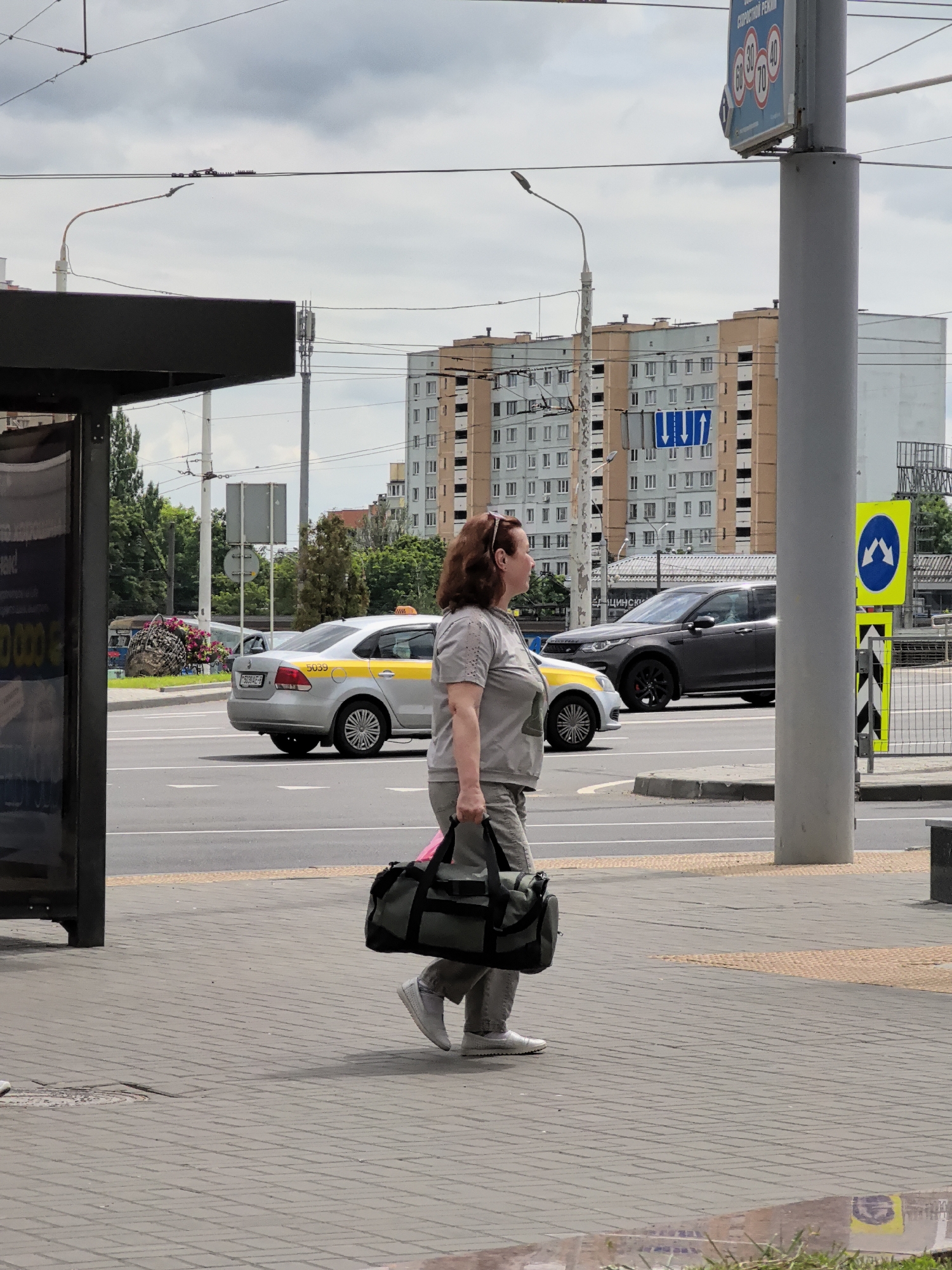 A medium shot captures a woman in a grey t-shirt and grey pants walking on a sidewalk in front of a street. She carries a dark green duffel bag in her right hand and wears white sneakers. Her brown hair is tied back. Behind her, a silver taxi with a yellow stripe is driving on the road, followed by a dark grey SUV. In the background, apartment buildings are visible under a cloudy sky, with power lines and street signs in the foreground. The sidewalk is paved with grey bricks, and a yellow and grey striped pole stands to the right of the woman.