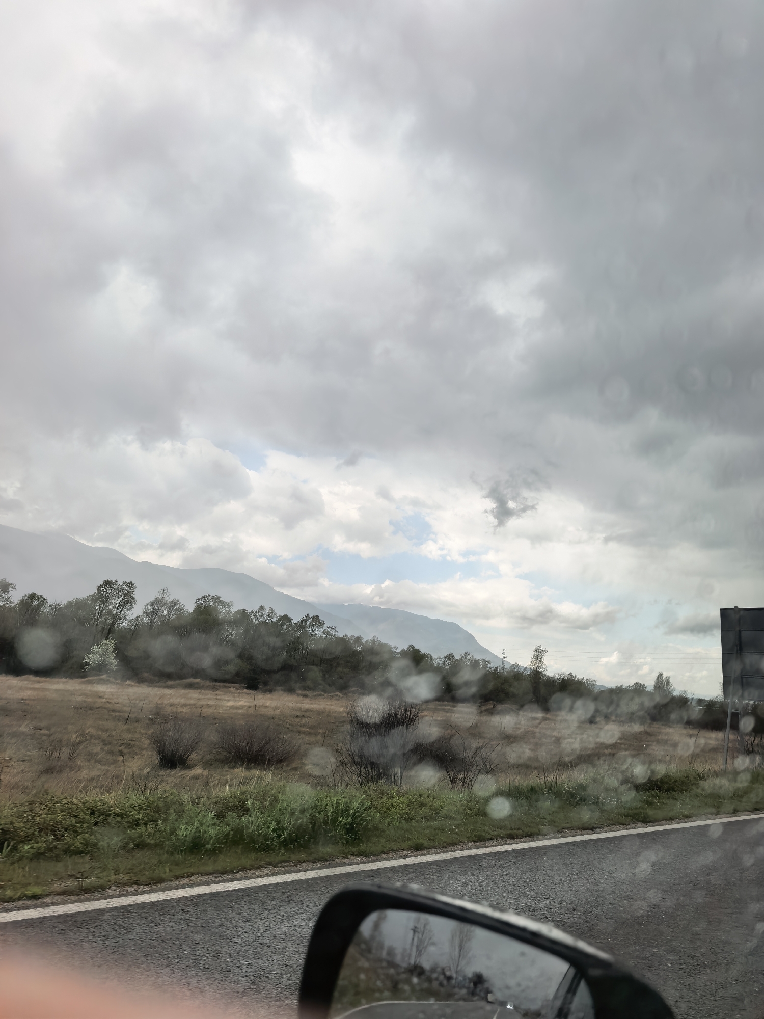 The image captures a vast, rural landscape viewed from inside a vehicle on a cloudy day. Raindrops are visible across the car window, indicating current or recent precipitation.
The sky is largely overcast with dense, grey clouds, though patches of lighter blue sky are visible towards the horizon, suggesting the clouds might be breaking. In the distance, a range of mountains appears hazy and muted under the cloudy sky. Below the mountains, a line of deciduous trees, some with early spring foliage or blossom, creates a demarcation line across the mid-ground.
The landscape transitions into expansive fields and scrubland, characterized by dry, yellowish grasses and scattered bushes. Closer to the foreground, a strip of greener vegetation runs alongside the road. An asphalt road with a white dividing line occupies the bottom portion of the image. The reflection in a car's side mirror is visible in the bottom right corner, showing a reversed view of the road and surrounding landscape. A person's arm or hand is partially visible in the lower left, further confirming the perspective from inside a car. A dark, rectangular structure, possibly a sign or part of a building, can be faintly seen on the far right edge of the frame.
The scene conveys a sense of travel through a quiet, natural environment under unsettled weather conditions. No discernible text is visible.