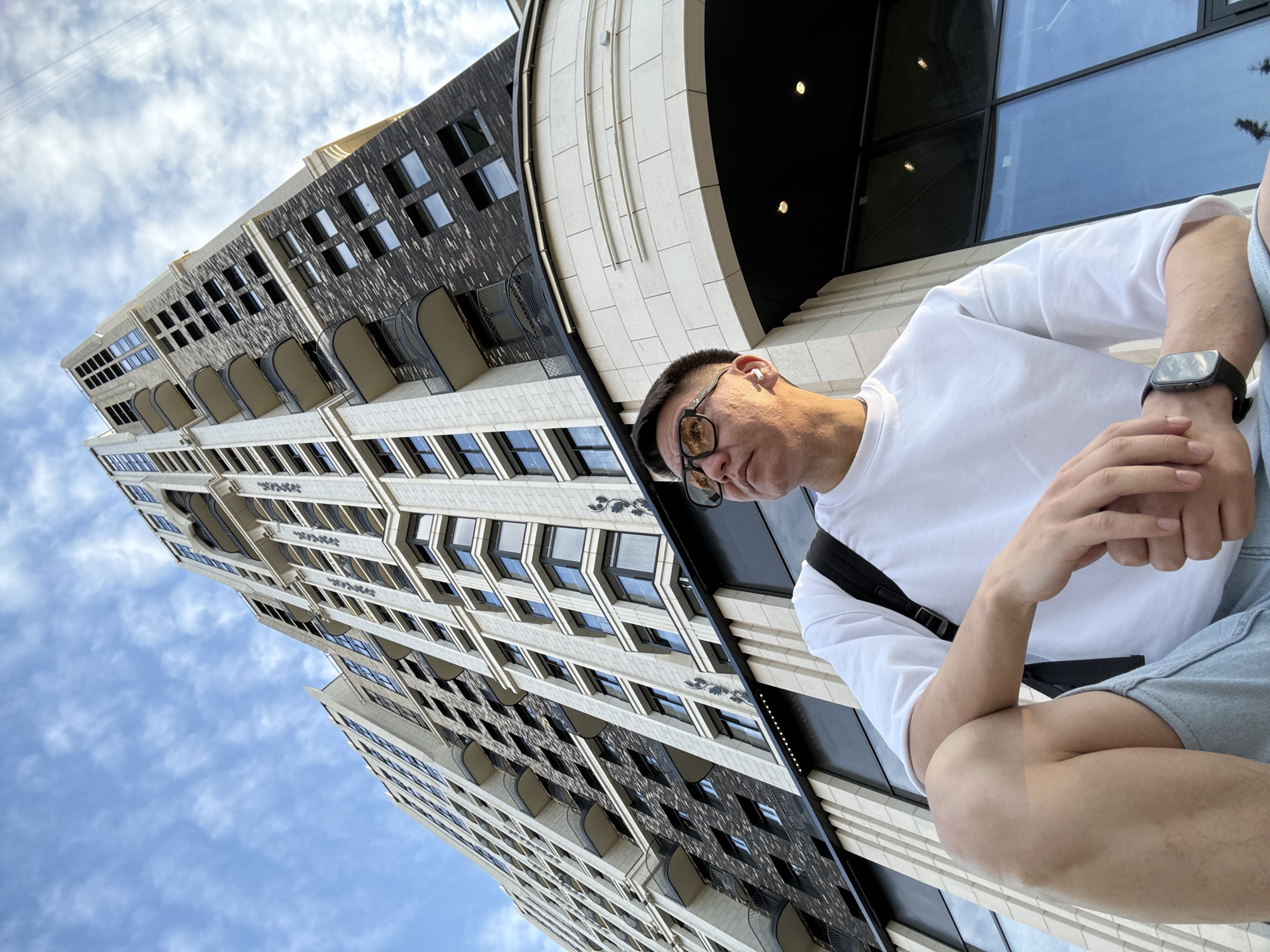 In Moscow, Russia, a low-angle outdoor shot features a man with short dark hair and clear skin wearing glasses, a white t-shirt, and light blue denim shorts. He has a black backpack slung over his left shoulder and a smartwatch on his left wrist. He is seated, with his left arm resting on his knee. Behind him, a modern high-rise building with a mix of white stone and dark brick facade towers into a partly cloudy blue sky. The building features numerous windows and balconies, and a large arched entranceway is visible in the foreground. The perspective emphasizes the scale of the building relative to the man.