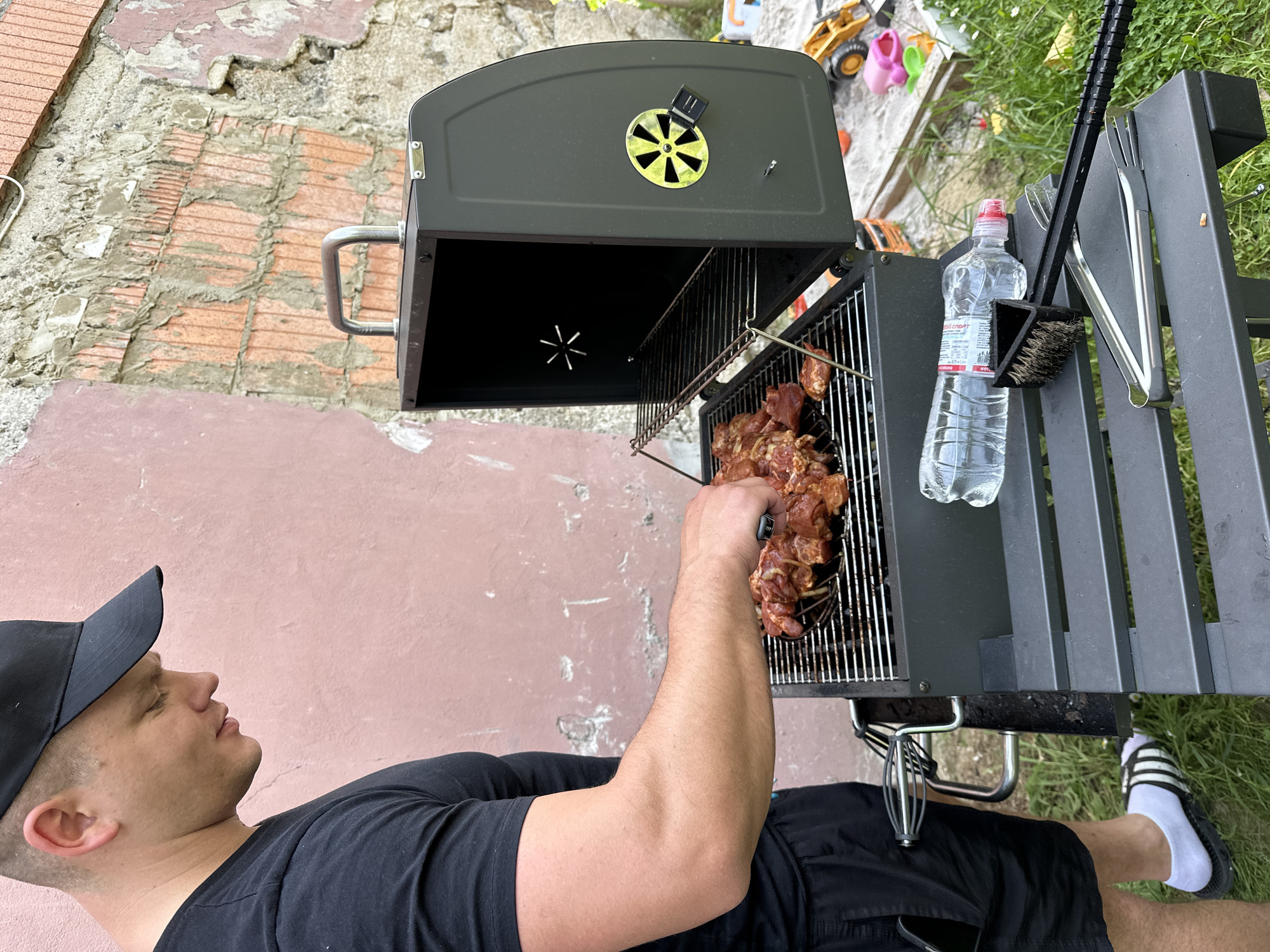 The image depicts a person grilling meat, presumably barbecue, on a portable grill located outdoors. The grill is black with a lid, and cooking utensils and a water bottle are nearby. The person is wearing a black t-shirt, black shorts, a black cap, and socks with sandals. The surroundings suggest a backyard or patio setting with brick and grass visible. The location is Niederquembach, Germany.