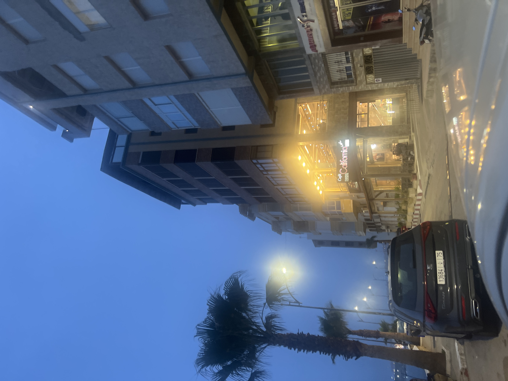 The image depicts a street scene in Fnideq, Morocco. A grey Seat Tarraco SUV is parked on the side of the road, its rear facing the camera. Its license plate reads "13684 75". Palm trees line the street on the left. The street is illuminated by streetlights. Buildings, some brightly lit, flank the street. A sign reading "Café California" is visible above a storefront. The sky is a pale blue. A car is partially visible on the right side of the frame.
