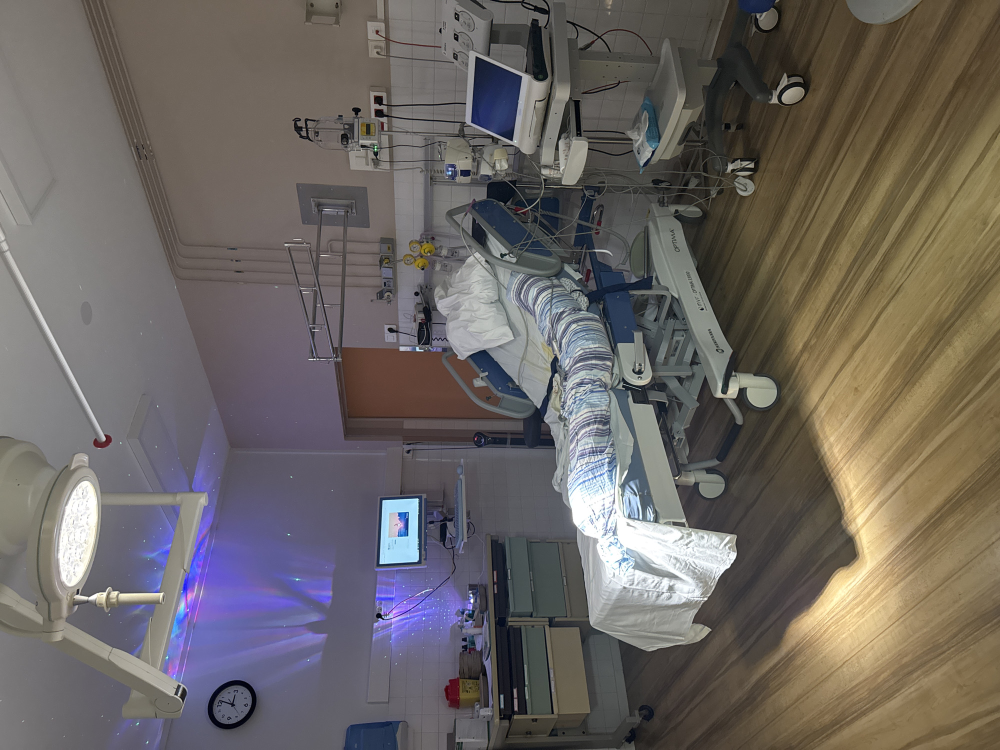 This is an image of a hospital room in Tourcoing, France. The room is equipped with a hospital bed, medical equipment such as a monitor, IV stand, and various devices connected by tubes and wires. The bed is made up with striped blankets and a white sheet. A wheeled medical cart with drawers is positioned to the left of the bed, and another wheeled cart with a monitor and medical supplies is to the right. The room has tiled walls and a wooden floor. A surgical lamp with colored lights is suspended from the ceiling, projecting patterns onto the wall. A clock is visible on the wall to the left. The overall scene depicts a clinical environment with a focus on patient care and medical technology.