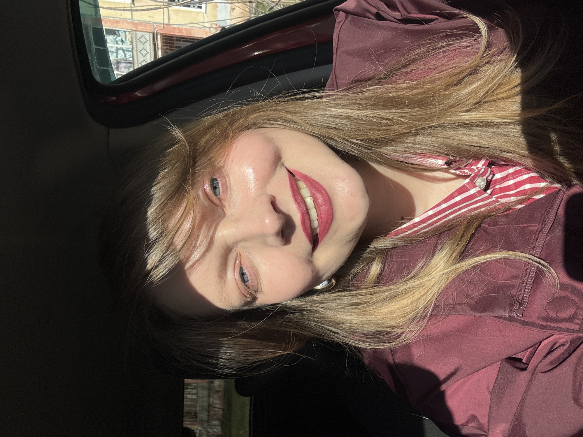 A light-skinned female with blue eyes and blonde hair is sitting in a car, smiling at the camera. She is wearing a burgundy jacket over a red and white striped shirt. The sun is shining on her face. The background visible through the car window shows urban buildings, suggesting a city setting. The location is Izhevsk, Russia, as indicated in the prompt.