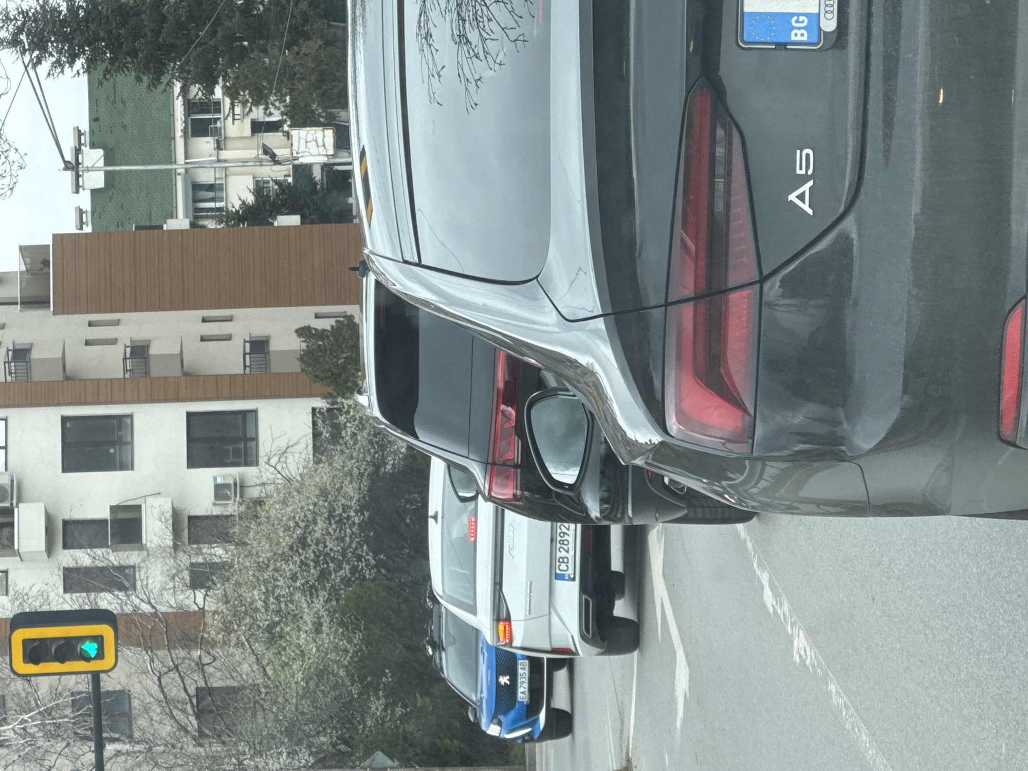 The image depicts a street scene in Sofia, Bulgaria, showing the rear of three cars waiting at a traffic light. The lead car is a dark grey Audi A5 with a Bulgarian license plate. Behind it is a white vehicle, possibly a Peugeot, with the license plate "CB 2892." The third car, barely visible, is blue and has the license plate "EA7935AB". The traffic light is green. Buildings are visible in the background, including a white building with visible air conditioning units and a brown building with multiple windows. The street appears to be an urban environment, with road markings.
