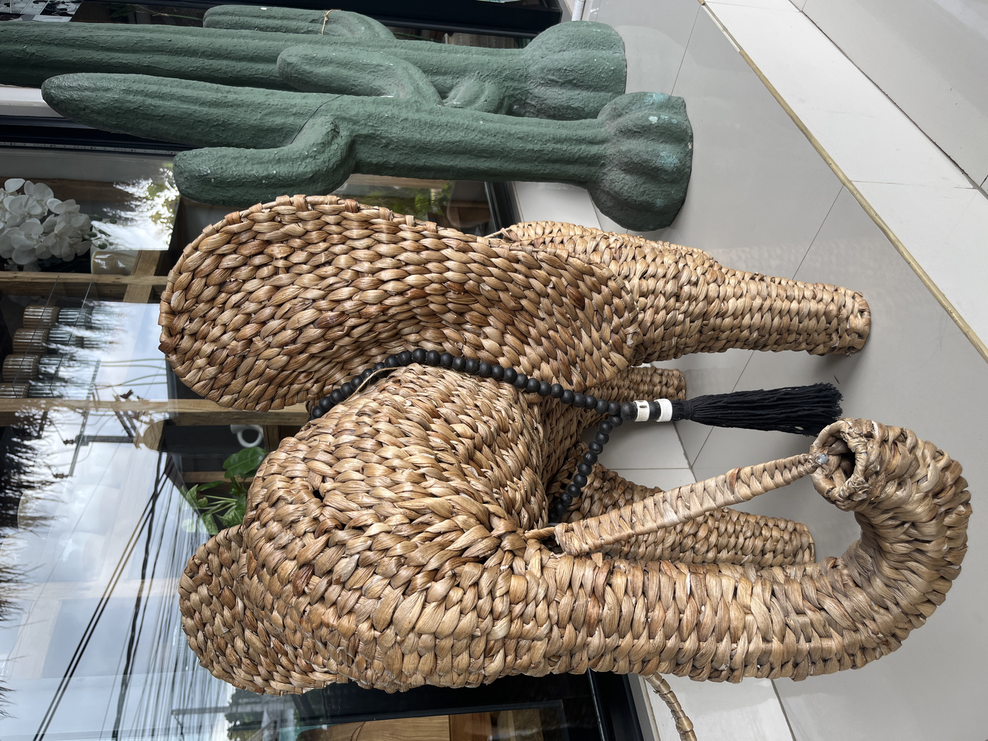 A woven wicker elephant figurine stands on a tiled ledge outside a storefront in Kuta Selatan, Indonesia. The elephant is a light brown wicker with a long trunk curled downwards. It has large, flat ears and a beaded black necklace with a black tassel hanging from it. Behind the elephant, two green, cactus-like sculptures stand. The storefront window reflects the sky and some power lines. The ledge is white with a thin gold trim.