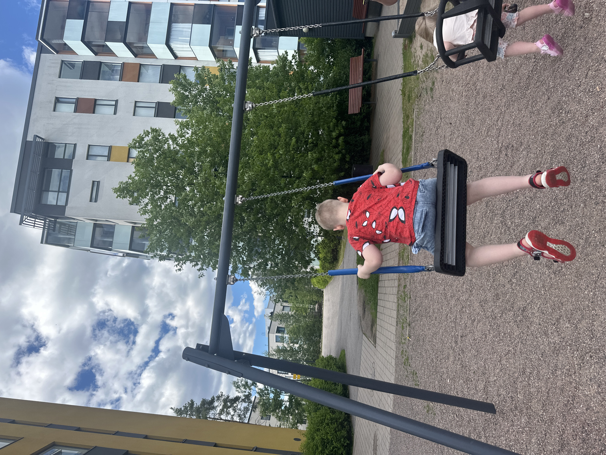 An outdoor, medium-shot, eye-level photograph shows a young boy and a young girl on swings in a playground. The boy, in the foreground, faces away from the camera. He has short brown hair and is wearing a red t-shirt with a white spider-web pattern and light blue denim shorts. He is wearing red sandals. The girl is on the swing to the right, also facing away from the camera. She has blonde hair and is wearing a white t-shirt and patterned shorts, and pink sandals. The swings are black with blue seats and chains. They are suspended from a dark grey metal A-frame structure. The ground is covered with small grey gravel. In the background, to the left, is a tall, yellow apartment building. To the right and center, behind the swings, are modern, multi-story apartment buildings with white and grey facades and balconies with glass and blue accents. Lush green trees and bushes are scattered throughout the scene, and a brown wooden bench is visible in the middle distance. The sky is cloudy with patches of blue.