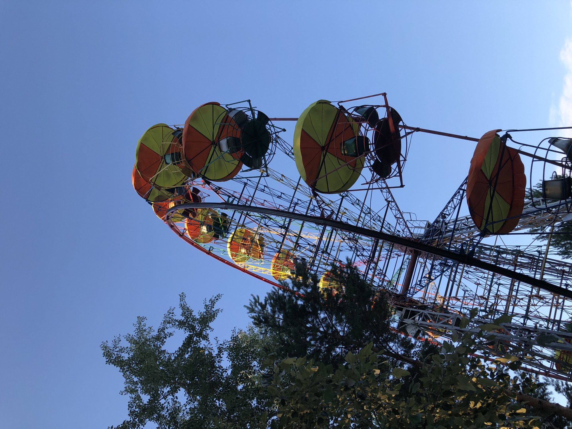 The image shows a Ferris wheel viewed from below against a clear blue sky. The Ferris wheel has a complex metal structure and is decorated with colorful gondolas. Some of the gondolas are visible, with orange and yellow striped canopies. The Ferris wheel appears to be illuminated with colorful lights, suggesting it might be evening or dusk, or that the lights are on even during the day. Trees with green foliage are visible in the foreground, partially obscuring the base of the Ferris wheel and some of its lower sections. The image captures the grandeur of the amusement park ride against the vastness of the sky, with the trees providing a natural element to the scene. The overall impression is one of a recreational setting, likely an amusement park or fairground. There is no visible text in the image. The setting is Chaikovsky, Russia.