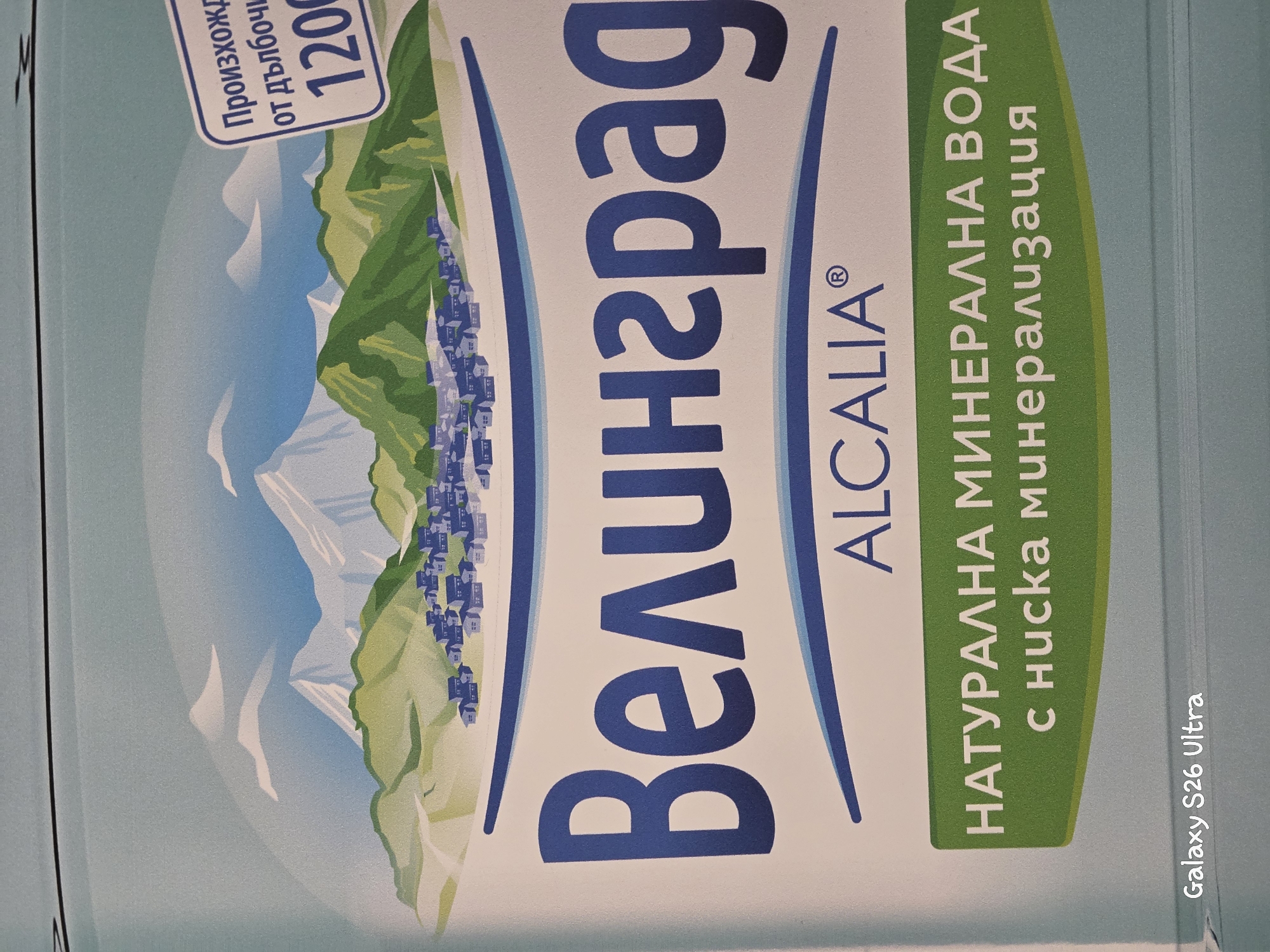 This is a close-up, rotated photograph of a product label for "Велинград" bottled water. The label features a scenic illustration of a mountainous landscape with a village nestled on a hillside. The water is described as "НАТУРАЛНА МИНЕРАЛНА ВОДА с ниска минерализация," which translates to "NATURAL MINERAL WATER with low mineralization." There is also text indicating that the water originates "от дълбочи," meaning "from the depths," and the number "1200" is prominently displayed, possibly indicating its depth of origin or a batch number. The brand name "ALCALIA®" is also visible. The overall aesthetic is natural and refreshing, aiming to evoke the purity and origin of the water. A "Galaxy S26 Ultra" watermark is present in the bottom right corner, indicating the device used for capture. The image's orientation suggests it might be a side panel of the water bottle or packaging.