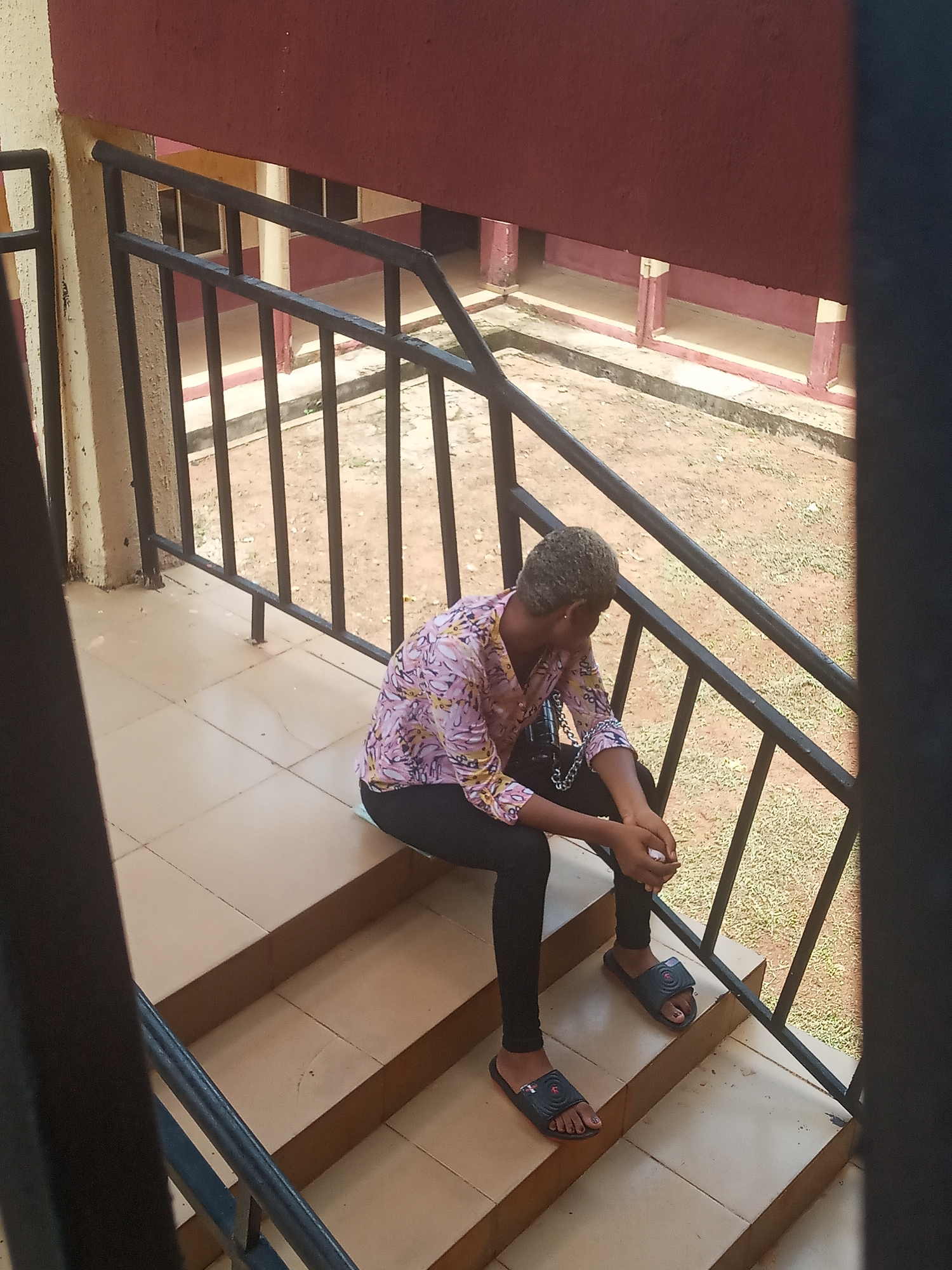 The image depicts a young woman sitting on an outdoor concrete staircase, viewed from a slightly elevated perspective. She has short, light-colored hair and is dressed in a long-sleeved shirt with a pink and yellow floral or abstract pattern, black leggings, and dark slides/sandals. Her head is gently bowed, and her hands are clasped in her lap, conveying a sense of contemplation, rest, or possibly introspection. A small chain is visible near her left arm, and a white object rests beside her on the stairs.
A dark metal railing runs parallel to the staircase, acting as a barrier and a visual frame. Beyond the railing, the ground is unpaved, showing dry soil and sparse patches of green grass. In the background, sections of a building or structure are visible, characterized by deep red walls and lighter, possibly beige, architectural trim or columns. The natural lighting suggests it is daytime, likely a dry and warm day, fitting the outdoor setting in Nkwelle, Nigeria. There is no visible text in the image.