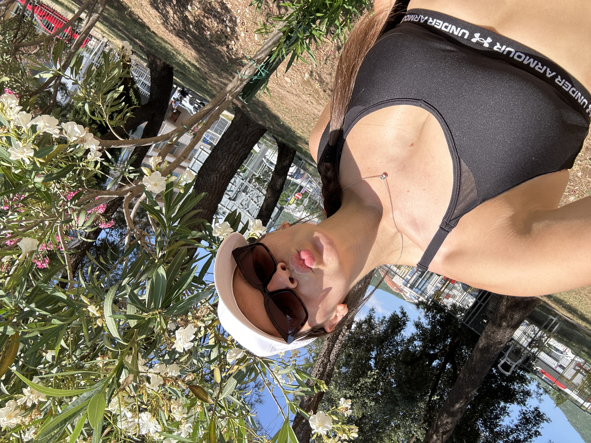 A woman in a white baseball cap and sunglasses is taking a selfie outdoors, with flowering trees in the foreground. She is wearing a black Under Armour sports bra and a necklace with a small diamond pendant. The background shows a grassy area with trees and buildings. The scene appears to be in Bečići, Montenegro, as evidenced by the architecture and vegetation typical of the region.