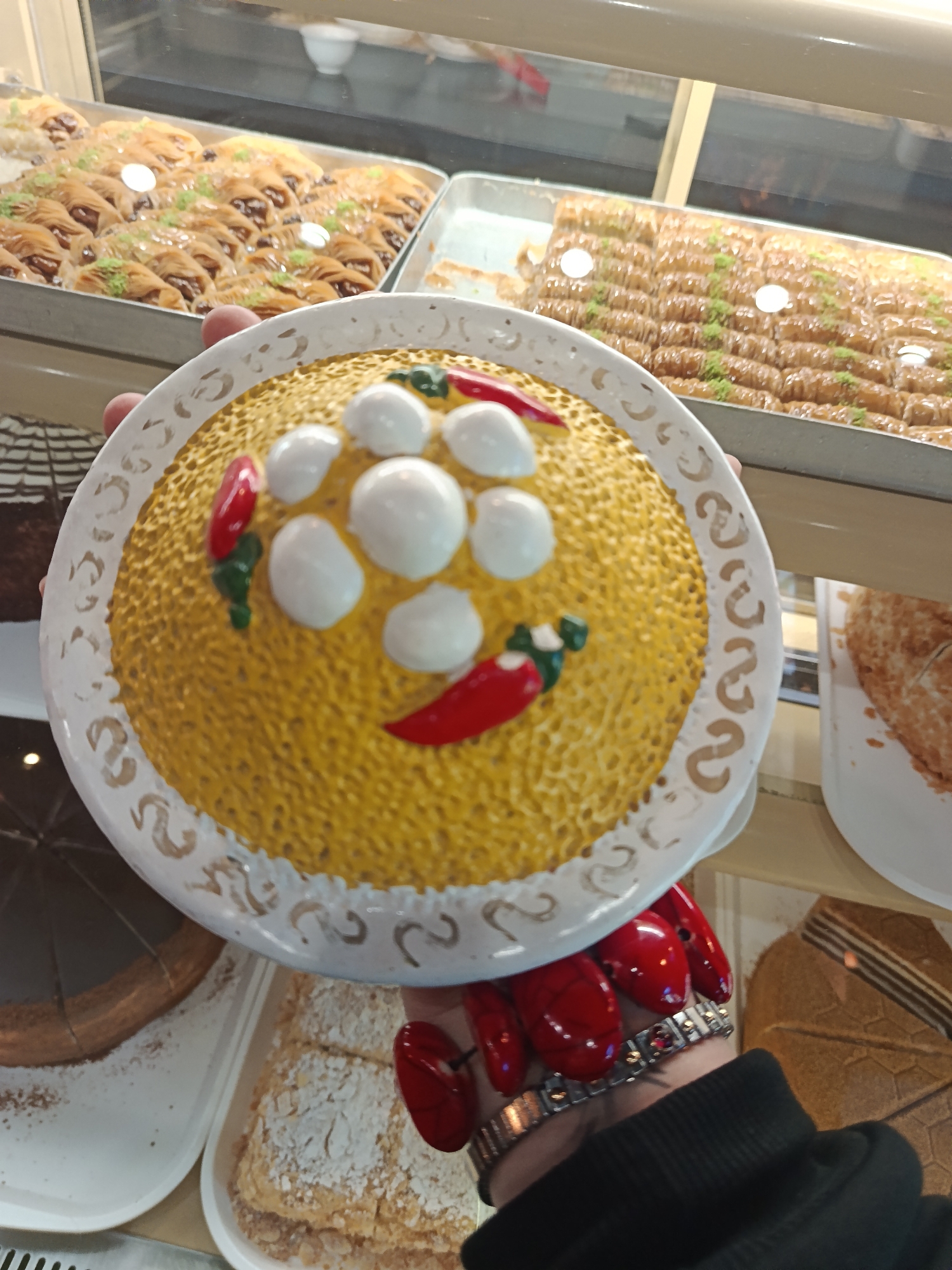 A close-up shot captures a plate of yellow, grain-like food decorated with white spheres and red peppers, resembling a face. The plate is held by a hand with red polished fingernails, adorned with a silver watch. Background includes a display case filled with various types of pastries and desserts. The setting appears to be a bakery or pastry shop. Moscow, Russia.