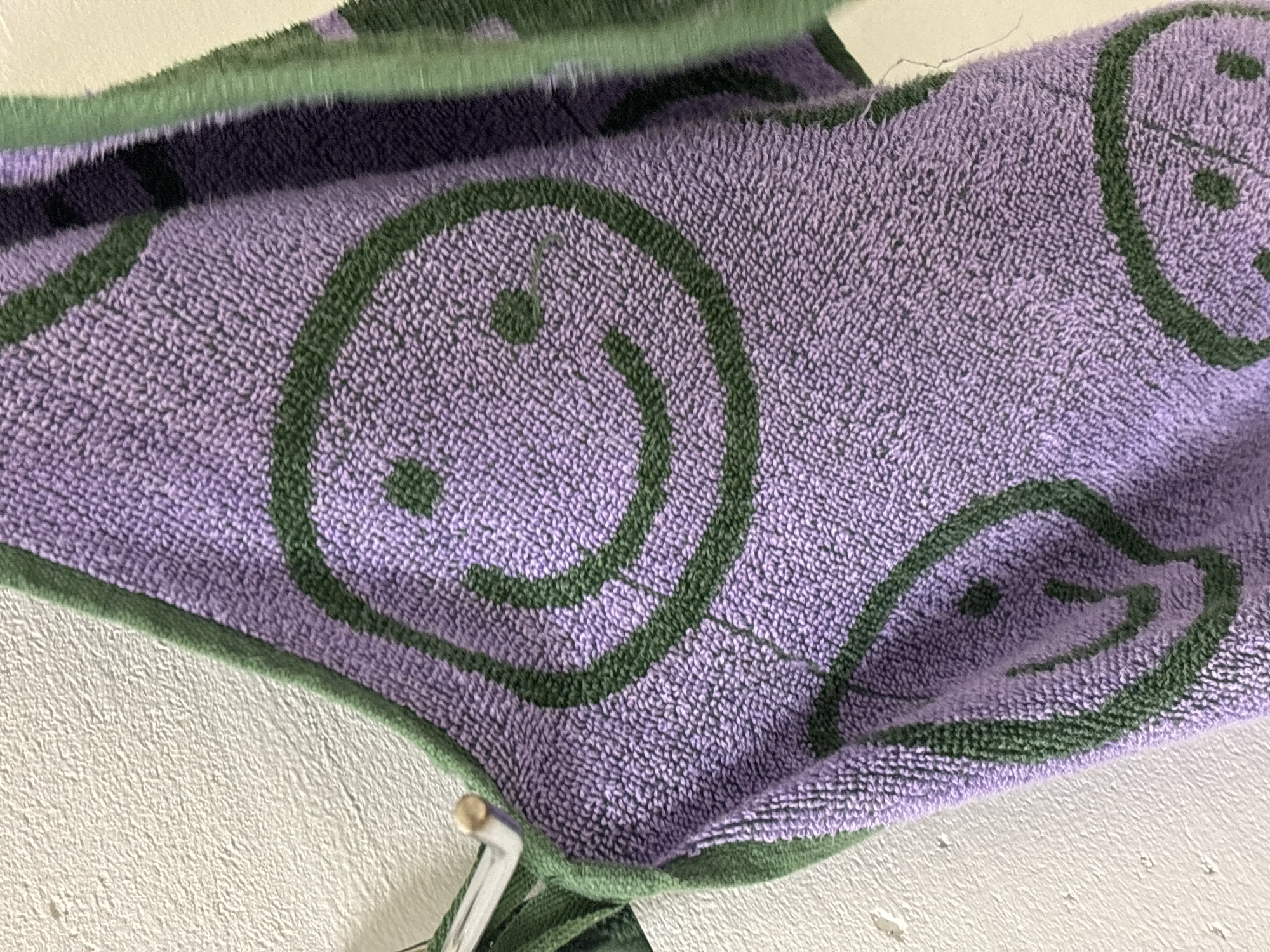The image is a close-up, high-angle shot of a purple towel with green smiley faces. The towel has a textured, terrycloth surface. It is draped over a white wall, which appears to be a rough, stucco-like texture. A metal hook with a small wooden knob is visible on the left side of the image, from which the towel is hanging. The lighting is soft and diffused. The scene is indoors, likely in a bathroom or laundry area, against a plain white wall. The location in the city of Ostrov, Russia is not discernible from the image content.
