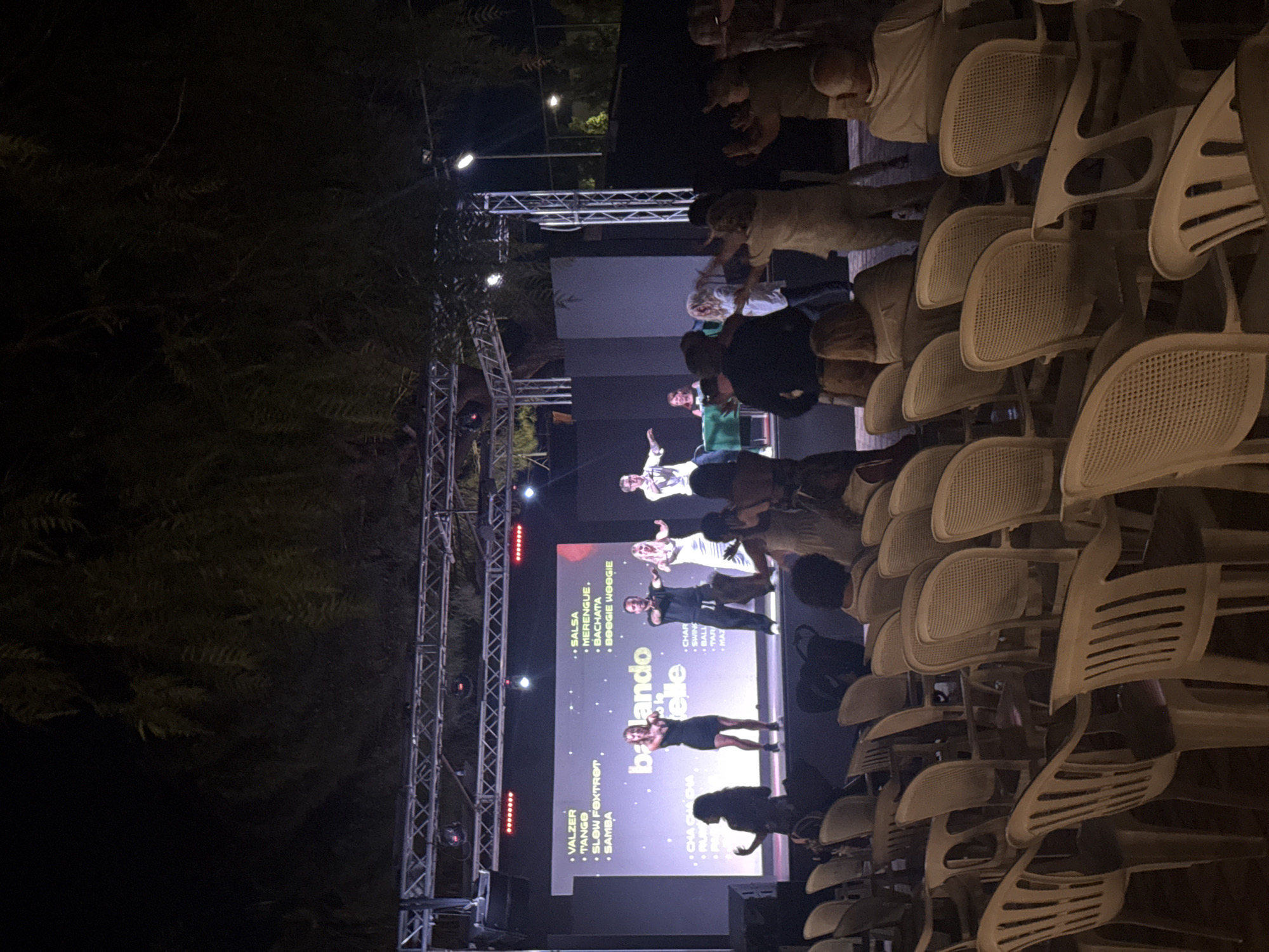 The image depicts an outdoor evening performance at what appears to be an event space in Isola delle Femmine, Italy, likely a piazza or an open-air venue. A stage is set up with a large screen displaying text related to dance styles such as "VALZER," "TANGO," "SALSA," "MERENGUE," "BACHATA," and "BOOGIE WOOGIE," with the words "balando le stelle" prominently featured. On the stage, several performers are engaged in dancing. One woman in a black dress is center-stage, and beside her, a man in a dark outfit dances with a blonde woman in a white dress. Further to the right, another couple dances. In front of the stage, an audience is seated in rows of white plastic chairs. Some audience members are visible in silhouette on the left, and others are more clearly seen in the foreground and mid-ground, watching the performance. The environment is dark, illuminated by stage lighting and some ambient light from the surroundings, with trees partially obscuring the top of the frame. The overall scene suggests a lively dance event or recital.