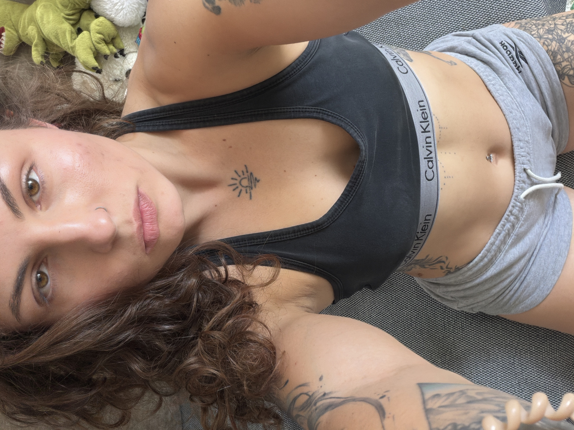 A young adult woman is lying on a grey textured surface. She wears a black Calvin Klein sports bra and grey Reebok shorts. She has curly brown hair, a nose piercing, and several tattoos visible on her chest, abdomen, and arms. Some stuffed animals are visible in the background. The location in Kiryat Bialik, Israel is not evident from the image.