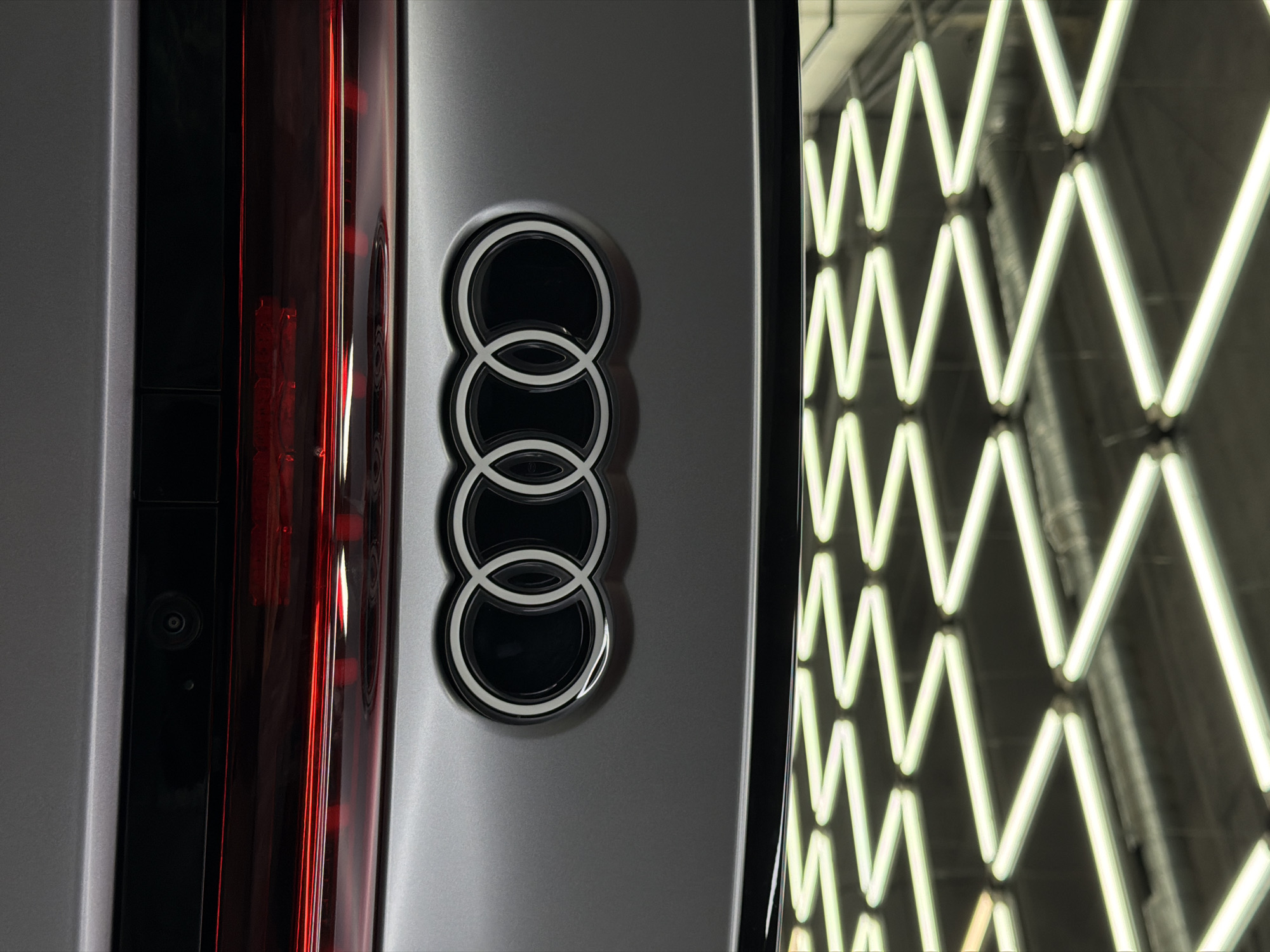 A close-up, vertical shot captures the rear portion of an Audi vehicle, likely indoors in a setting such as a showroom or dealership in Krasnoyarsk, Russia. The primary subject is the iconic Audi logo, featuring four interlocking rings, prominently displayed on a sleek, matte dark gray panel of the car. The rings themselves are rendered in a metallic silver or white, outlined with a distinct darker border, creating a sophisticated contrast.
To the left of the Audi logo, a vertical, dark red taillight or brake light assembly is visible, reflecting a deep, glossy sheen. Below the upper part of this taillight, a small, circular camera lens, likely for a rear or side-view system, is subtly integrated into the car's bodywork. The vehicle's panel has a smooth, refined texture.
The right side of the image is dominated by the reflection of bright, white, zig-zag patterned light tubes, which appear to be part of a wall or ceiling lighting fixture in the environment. These lights create a dynamic, modern backdrop, albeit slightly out of focus, adding depth and suggesting a contemporary, well-lit space. No people are visible, and no overt activity is taking place beyond the static display of the car's details. The image emphasizes the brand's aesthetics and technological integration.