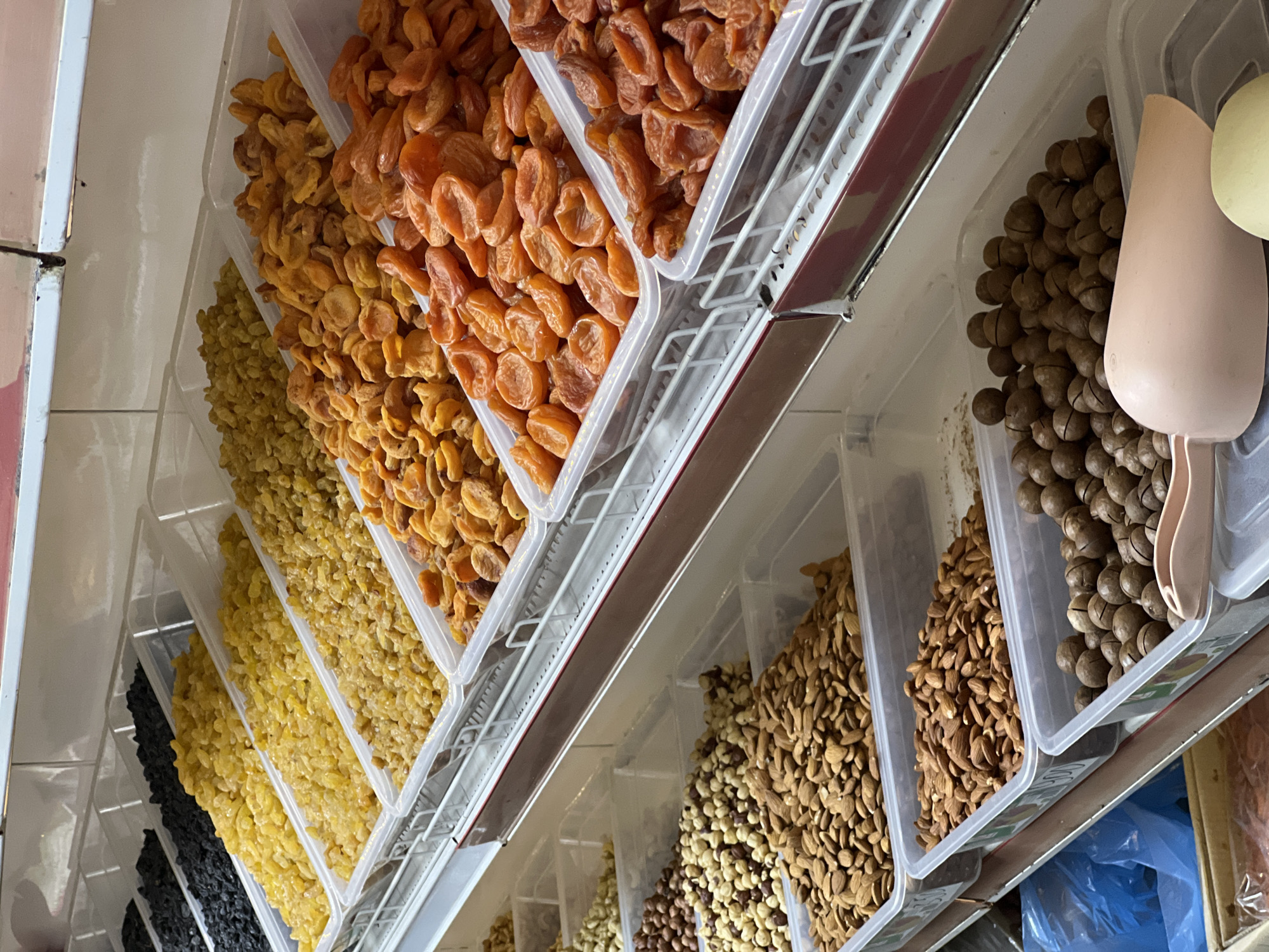 The image shows a variety of dried fruits and nuts displayed in clear plastic containers, arranged in rows on shelves, likely in a market or shop. From left to right, the visible containers contain: dried black currants, yellow raisins, golden raisins, dried apricots, another type of dried apricot with an orange hue, and then a variety of nuts, including almonds, walnuts, macadamia nuts, and hazelnuts. A couple of light-colored plastic scoops are partially visible in the rightmost container. The setting is indoors, possibly a shop in a city, but it's not possible to determine the location as Krasnodar, Russia from the image alone.