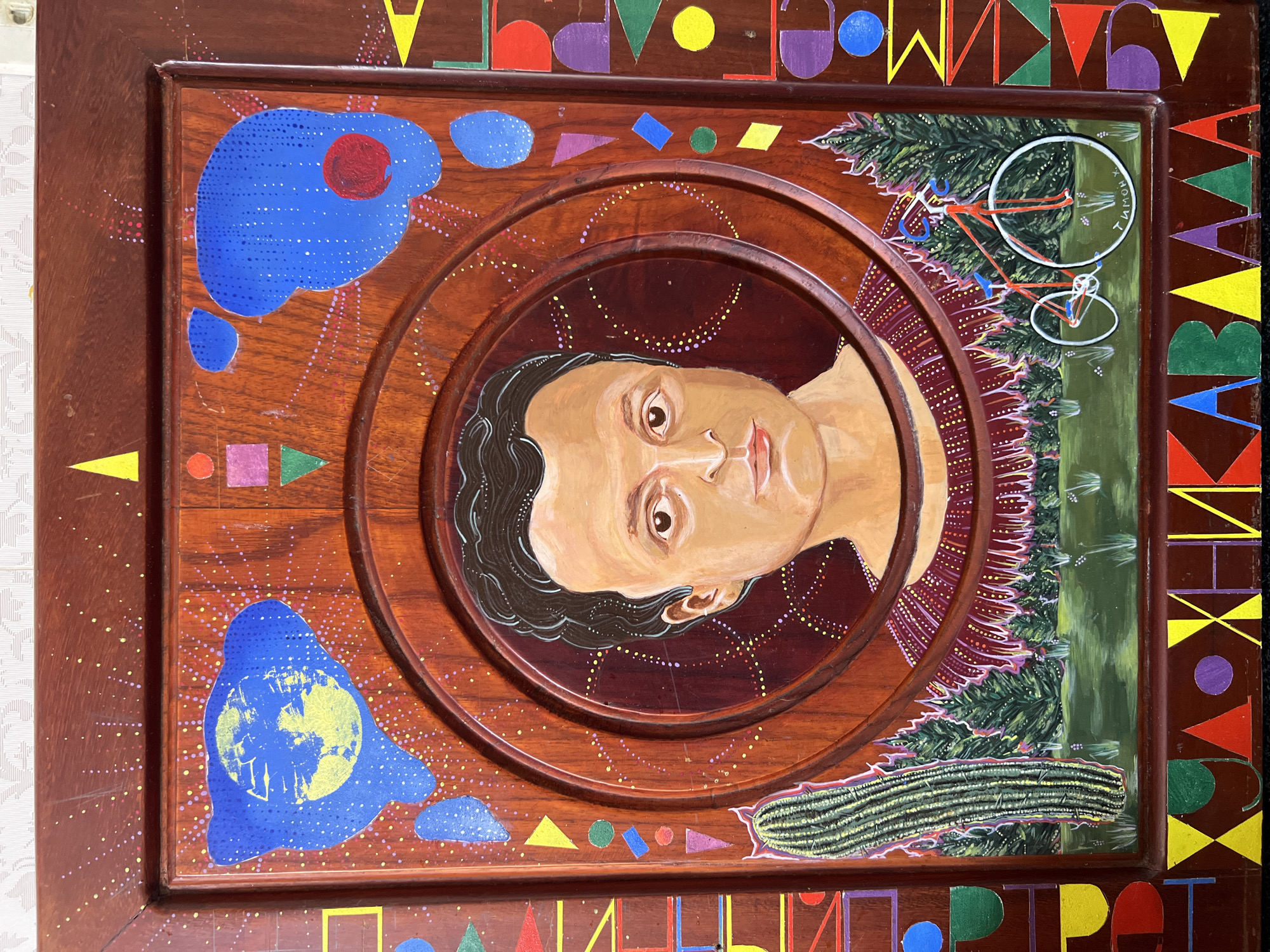 This image captures a vertical, mixed-media artwork, likely a painted wooden panel or door, displayed indoors against a light-colored, textured wall. A small white electrical box or hook is visible on the wall in the upper right corner of the photograph's background. The artwork itself is located in an indoor setting within Nha Trang, Vietnam; no specific address or landmark is discernible from the image content.
The artwork features a stylized portrait of an individual with light skin, dark wavy hair, brown eyes, and a neutral expression, depicted from the upper chest. This portrait is centrally positioned within a series of concentric, raised wooden rings, forming a circular frame. The figure wears a dark red garment with a patterned texture around the neck and shoulders.
The background of the artwork is a reddish-brown wooden surface. Above the portrait, this background is scattered with numerous small yellow, pink, and white dots. Two irregular blue shapes are present; the one on the left contains a stylized yellow depiction of the Earth, while the one on the right contains a solid red circle. Below the portrait, a landscape scene is depicted with a row of dark green, conical trees bordering a green grassy foreground featuring small white and purple plants. A tall, ribbed green cactus stands vertically on the left side of this landscape. On the right, a red bicycle with white wheels and blue handlebars is depicted, standing in the grass. Near the bicycle, the Cyrillic script "ТИМОН Х." is visible.
The entire central painted area of the artwork is bordered by a wider reddish-brown wooden frame, which is decorated with a repeating pattern of colorful geometric shapes (triangles, squares, circles, rectangles) and stylized block letters. The visible letters along the bottom border include "ХНИКАВААА".