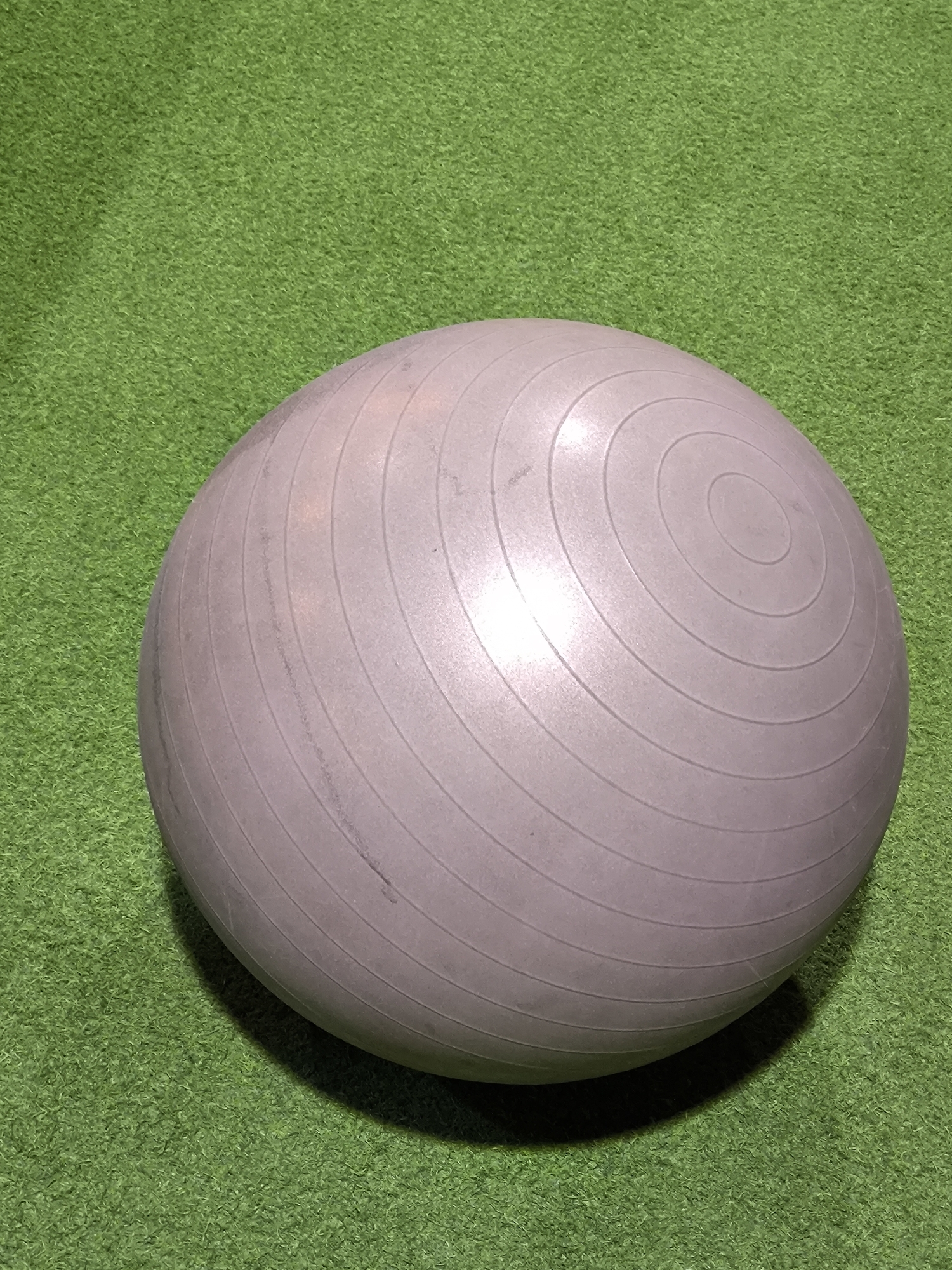 An exercise ball rests on artificial green turf in an indoor facility in the city of Unknown, Unknown. The ball is large, spherical, and grey with a matte finish. Concentric circular grooves are visible across its surface, particularly on the upper right side. Minor scuff marks and discoloration are present on the ball's surface. It casts a shadow directly beneath and behind it, indicating a light source from the front-right. The green turf has a uniform, short pile, resembling synthetic grass. No people, other subjects, or explicit actions are depicted.