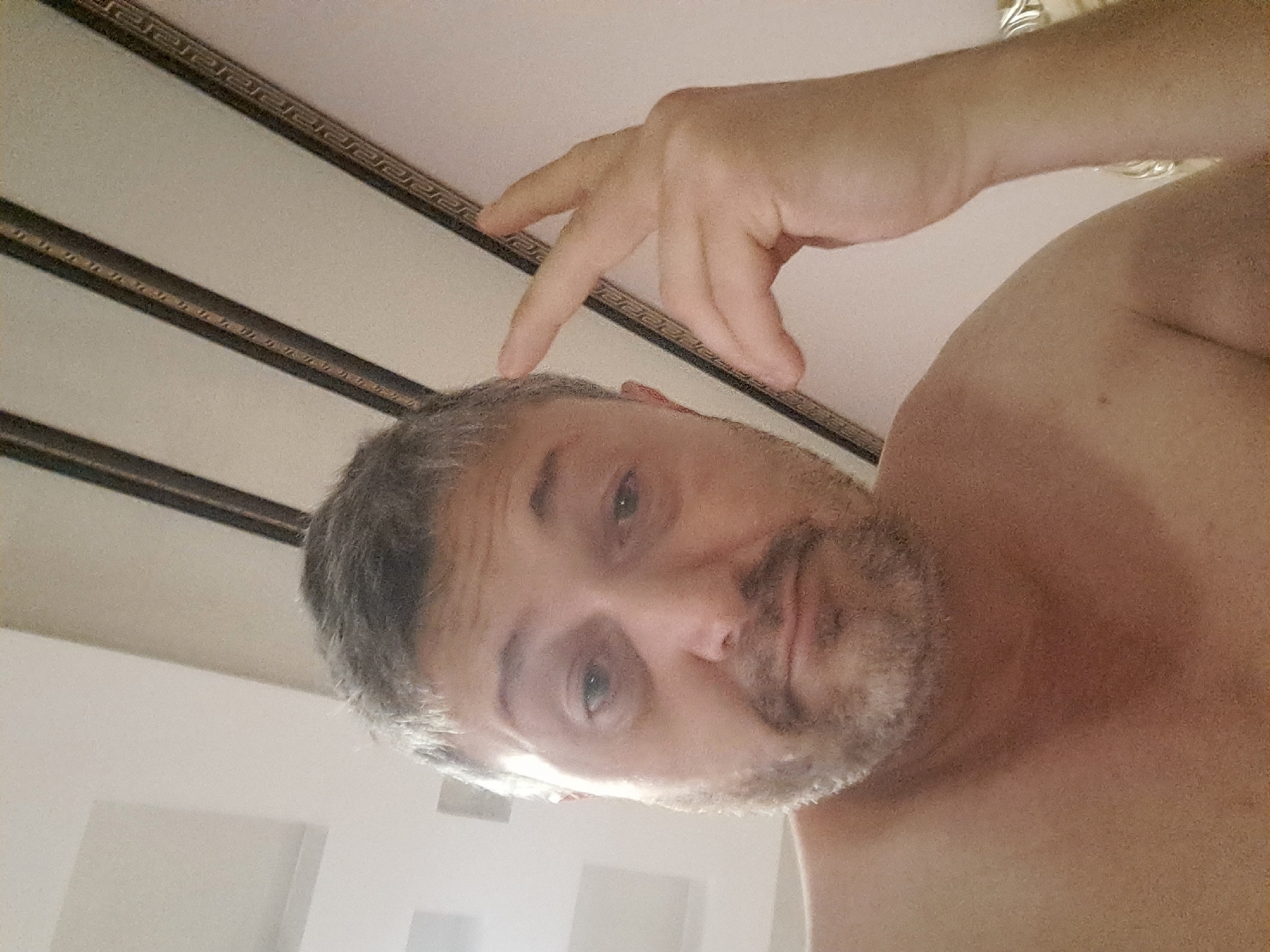 The image is a close-up selfie of a middle-aged man with graying hair and a beard, taken indoors. He is looking directly at the camera with a relaxed expression, and his right hand is raised to his temple, with two fingers extended. The man is shirtless, exposing his upper chest and shoulder. The background shows a ceiling with dark decorative beams and some recessed lighting fixtures. There are no discernible elements in the image that would place the location in Namangan City, Uzbekistan.