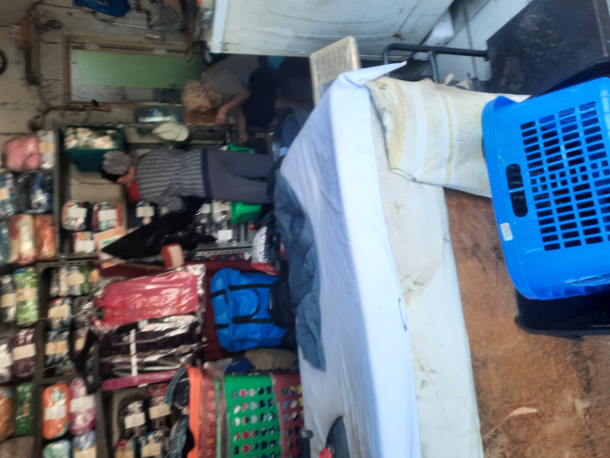 The image shows a cluttered indoor scene, likely a small shop or storage area. In the foreground, a mattress is covered with a light blue sheet and what appears to be dark grey fabric. A large, bright blue plastic basket with a grid pattern sits on top of a black scale in the lower right corner. To the left, there are stacked plastic baskets, one orange and one green, containing what look like folded items or packaging. Behind these, a large blue duffel bag stands upright.
In the background, shelves are laden with various items, many of which are packaged and appear to be textiles or clothing. A person wearing a striped shirt and dark pants is partially visible on the left, facing away from the camera and towards the shelves. Another person with curly hair, wearing a light-colored headscarf and a grey shirt, is seated on the right, appearing to be engaged in an activity. A doorway with a green door is visible in the background, leading to another space. The environment appears dimly lit and somewhat disorganized, suggesting a working or storage space. The presence of multiple baskets, packaged goods, and a scale points towards a retail or distribution context.
Without specific external information, it is impossible to definitively place this image in North Jakarta, Indonesia. However, the visual cues such as the types of goods, storage methods, and general aesthetic could be consistent with small commercial establishments found in urban areas of Indonesia.