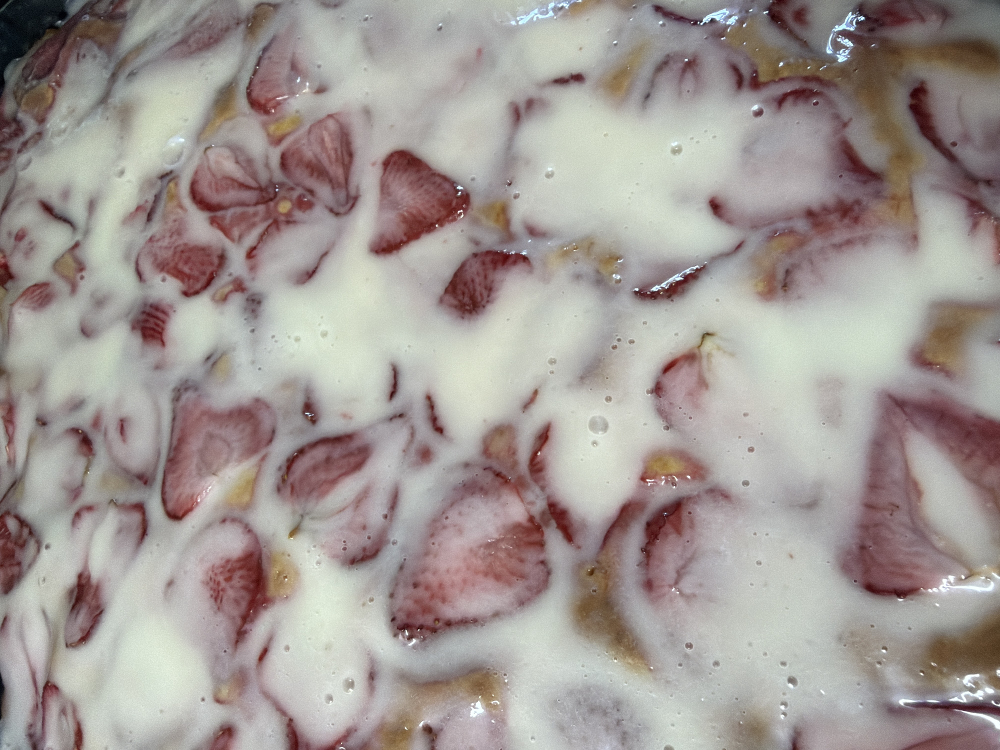 A close-up shot displays what appears to be a strawberry dessert, possibly a cake or tart, heavily covered with a creamy white topping. Sliced strawberries are embedded within the topping, revealing their red flesh and seeds. The base beneath the topping shows a brownish hue, suggesting a baked crust or cake layer. The cream is glossy and bubbly, indicating a recent application. The location of Chaikovsky, Russia, is not identifiable in this image.