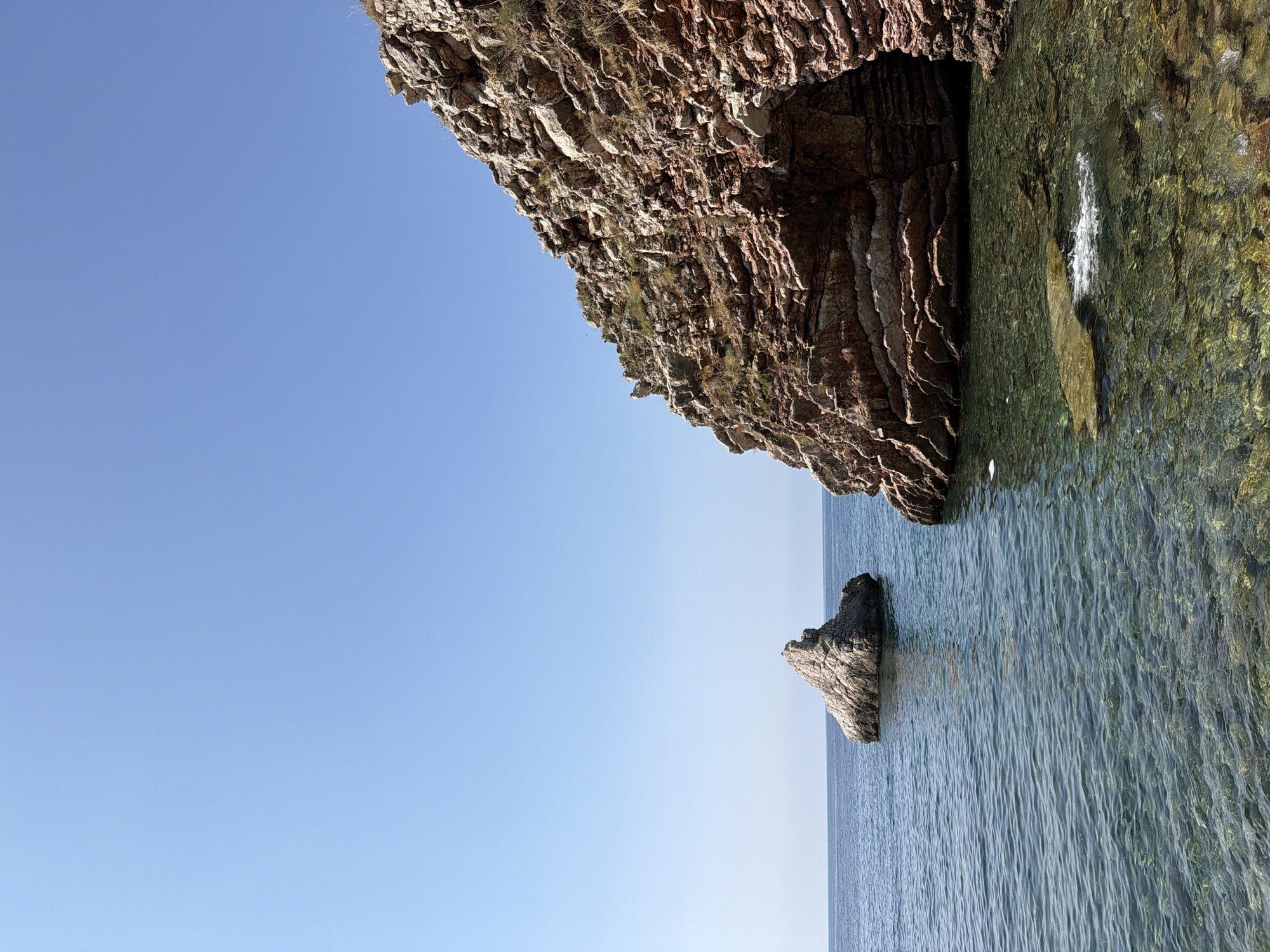 A vertical, medium shot captures a serene coastal scene under a clear blue sky, likely in Boreti, Montenegro. The foreground showcases the crystal-clear, turquoise waters of the Adriatic Sea, revealing a rocky seabed. The water laps gently against a rocky shoreline on the right, where a natural cave or archway is visible within a steep, layered cliff face. These cliffs, predominantly red and brown, are dotted with sparse vegetation. In the mid-ground, a single, large, grey rock formation rises dramatically from the sea. The horizon line is low, emphasizing the vastness of the sky. No people or animals are present in the image.