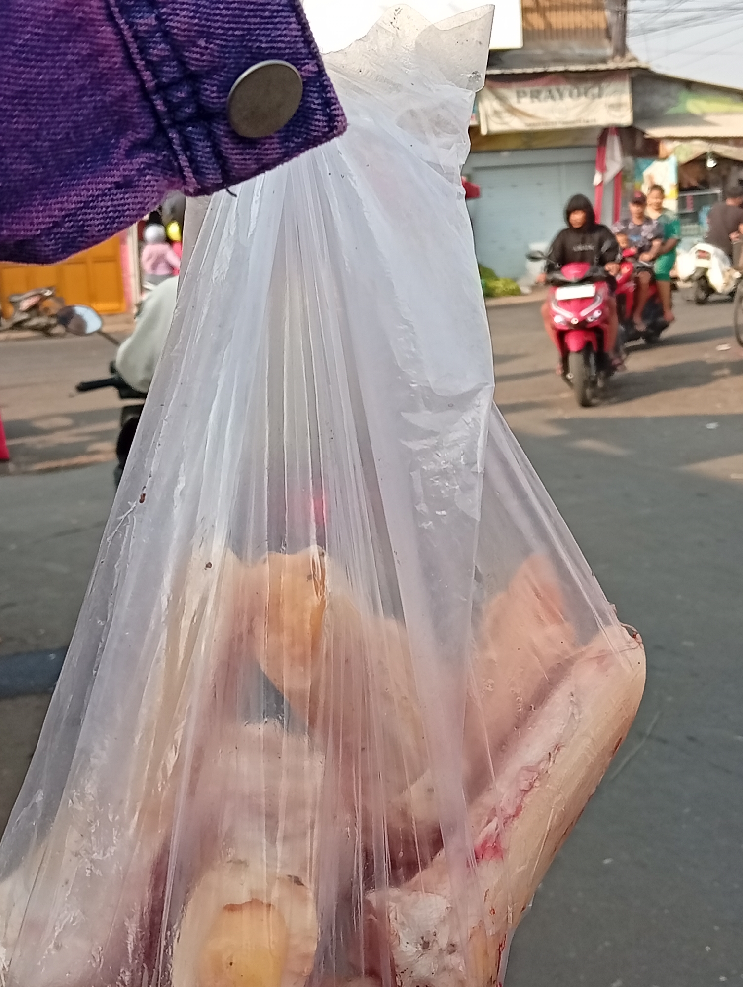 In the city of Larangan, Indonesia, a person holds a clear plastic bag filled with raw meat. The bag is partially transparent, revealing chunks of pale pink meat with some reddish-brown areas. The fabric of a purple garment, possibly a jacket, is visible at the top left, featuring a metal button. In the background, a street scene unfolds with several people on motorcycles. One individual in a black hoodie rides a red scooter towards the viewer. Other figures and a building with a banner reading "PRAYOGI" are also discernible. The overall environment appears to be a bustling urban street during daylight.