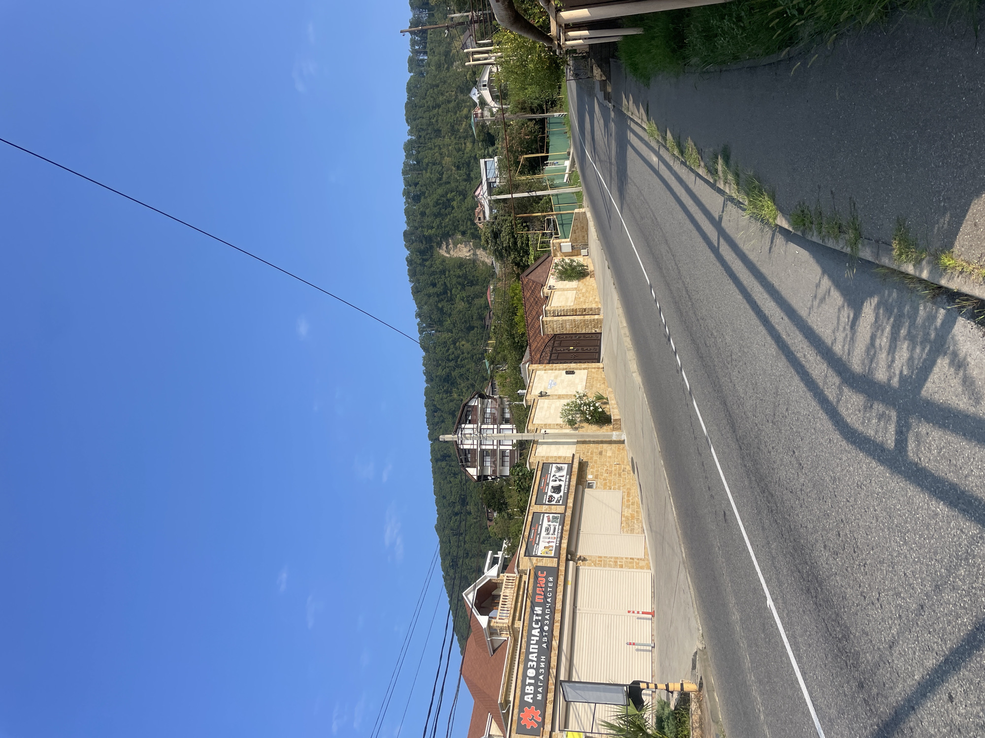 The image depicts a street scene in Baranovka, Russia. A paved road runs diagonally from the foreground to the background, with a white line marking the center. A sidewalk runs along the right side of the road, bordered by a metal railing and green grass. On the left side of the road, a building with a brown roof and light-colored brick facade is visible. The building features two large garage doors, one of which is partially open. A sign above the garage doors reads "АВТОЗАПЧАСТИ ПЛЮС" and "МАГАЗИН АВТОЗАПЧАСТЕЙ," indicating it is an auto parts store. Further down the street, more buildings and houses are situated on a hillside covered with dense green trees. The sky is clear and blue. The overall scene is a quiet, sunny day in a residential area with commercial elements.