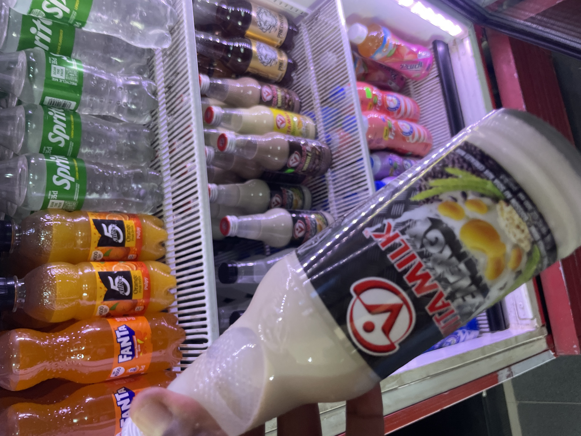 The image shows a refrigerated display case filled with various beverages in Kurunduma, Nigeria. On the left, there are bottles of Sprite, Fanta, and 5 Alive orange juice. In the middle, there are bottles of Predator energy drink and what appears to be various flavors of VITA MILK, including a bottle being held up in the foreground. In the background, there are bottles of peach-flavored drinks. The scene suggests a local store or market.