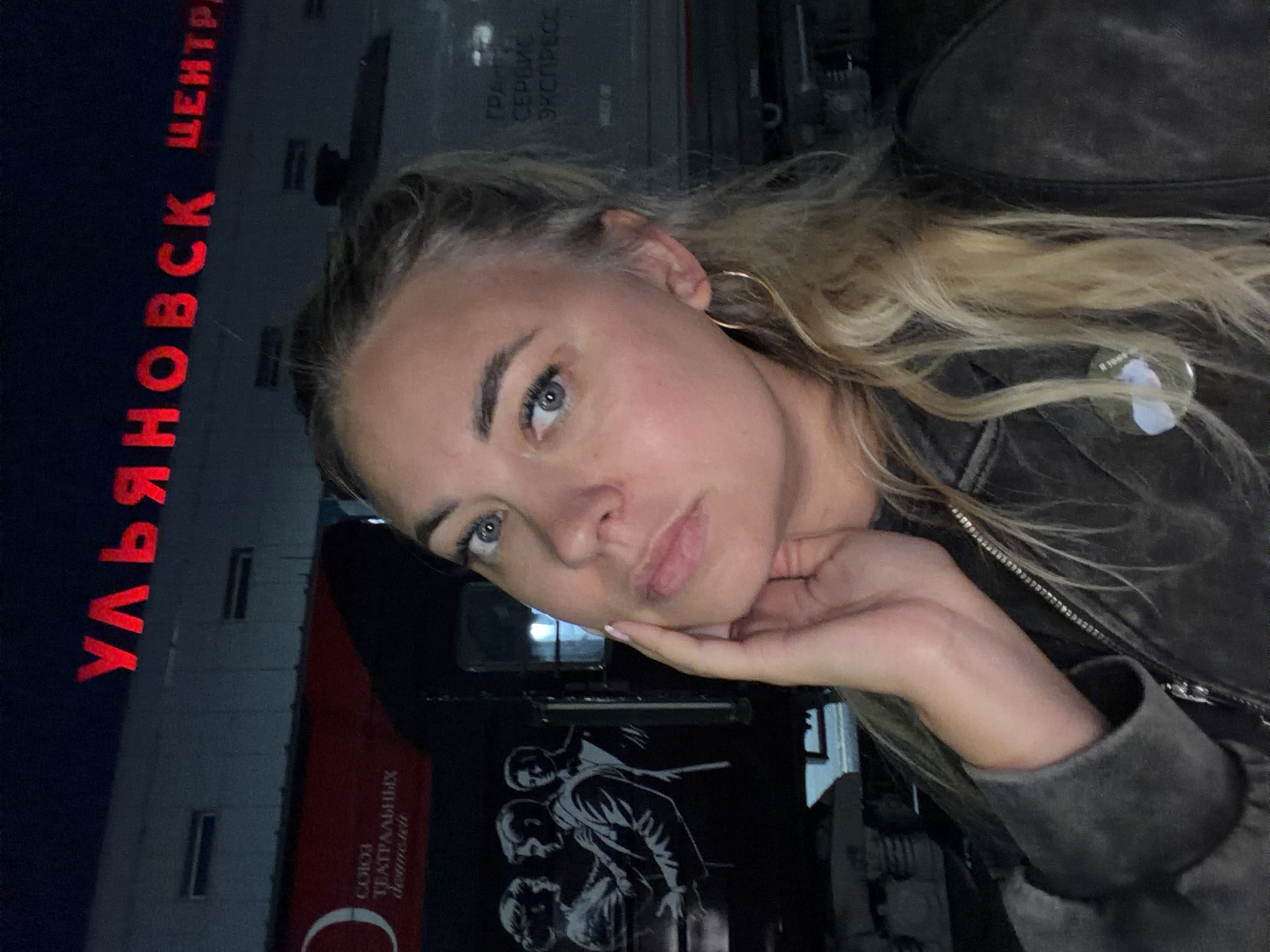 A medium shot depicts a young Caucasian woman with blonde hair, fair skin, and blue eyes, posing for a selfie at night in front of a building. She is wearing a dark, possibly denim or leather, jacket. Her left hand is raised to her cheek, and she is looking directly at the camera. Behind her, a building facade is visible, illuminated by red neon lights that spell out "УЛЬЯНОВСК ЦЕНТР" (Ulyanovsk Center) in Russian. To the left of the woman, a large red banner with white text is displayed on a dark surface, likely a train. The banner reads "СОЮЗ ТЕАТРАЛЬНЫХ ДЕЯТЕЛЕЙ" (Union of Theatre Workers). Below the banner, a graphic illustration depicts three silhouetted figures, two adult males and a female, appearing to be in a formal setting. The overall environment suggests an urban setting at night, possibly a train station or a cultural venue in Ulyanovsk, Russia.