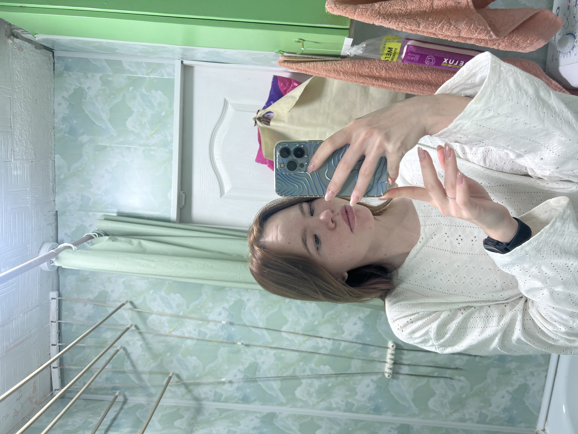 A fair-skinned person with reddish-brown hair takes a selfie in a bathroom. The person wears a white, eyelet-style blouse and a black fitness tracker on their left wrist. They are holding a blue iPhone in their right hand. The bathroom features green-toned tile walls, a green overhead trim, and a white door. A metal drying rack is visible on the left side of the frame. Towels and packaged goods are stacked behind the person near the top of the door. The setting is indoors and appears to be a personal residence. The image was taken in Chaikovsky, Russia.