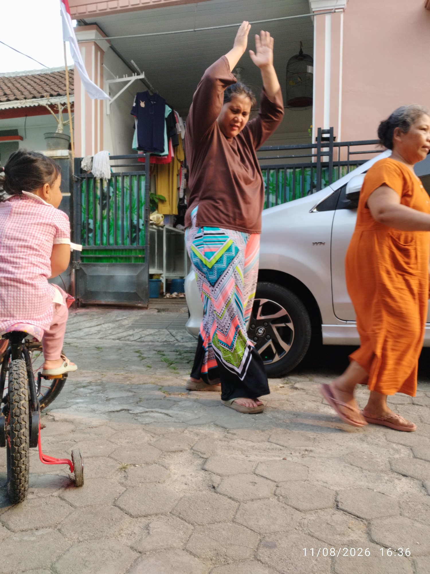 A side-view medium shot shows three females of various ages in front of a residential building. The youngest, a child, is seated on a bicycle with training wheels, facing left. A woman in a brown shirt and a patterned skirt stands in the middle, with her arms raised, while an older woman in an orange dress walks past on the right. A silver car is parked behind them. The scene is set on a paved courtyard with hexagonal stones, typical of a residential area in Bandar Lampung, Indonesia. A national flag hangs on a pole to the left of the frame.