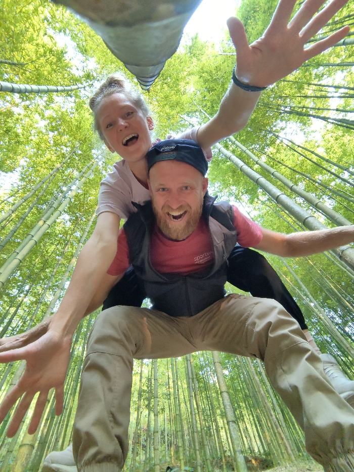 The image displays two adults, a male and a female, situated within a dense bamboo forest in Mù Cang Chải, Vietnam. The male, positioned centrally in the foreground and crouching, wears a backward dark blue baseball cap, a red t-shirt under a dark grey sleeveless vest, and tan pants. He has a light-colored beard and an open-mouthed smile, with his left arm extended laterally to the left. The female is positioned on the male's back or shoulders, wearing a light pink t-shirt with her hair pulled back. She also exhibits an open-mouthed smile and extends both arms laterally, with a dark bracelet visible on her right wrist. Both individuals are looking directly into the camera. The environment consists of numerous tall, slender, vibrant green bamboo culms and dense foliage, extending vertically into the bright upper frame.