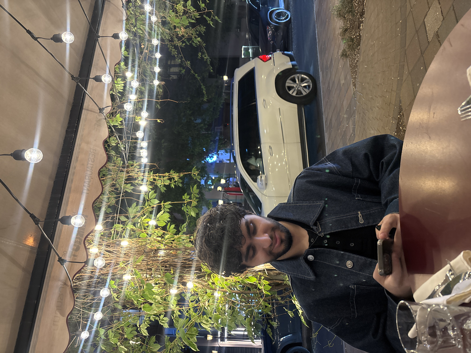 A man is seated at an outdoor table at a restaurant, holding a mobile phone. He is wearing a dark denim jacket over a dark shirt. The restaurant has a beige awning decorated with strings of lights and greenery. Behind him, through a window or opening, a white van and a dark car are parked along a street. The scene is located in the city of Yerevan, Armenia. The table is reddish-brown. There are glasses and tableware present.