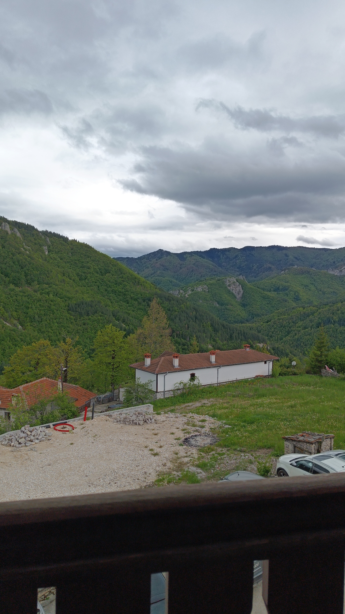 The image shows a mountainous landscape in Vrata, Bulgaria under a cloudy sky. In the midground, there's a white building with a red tile roof and chimneys, situated on a grassy area. To the left, a building with a similar roof sits near a gravel area with piles of rocks and a red object. The background features green, forested mountains. In the foreground, a dark wooden railing partially obscures the view, with a glimpse of vehicles.