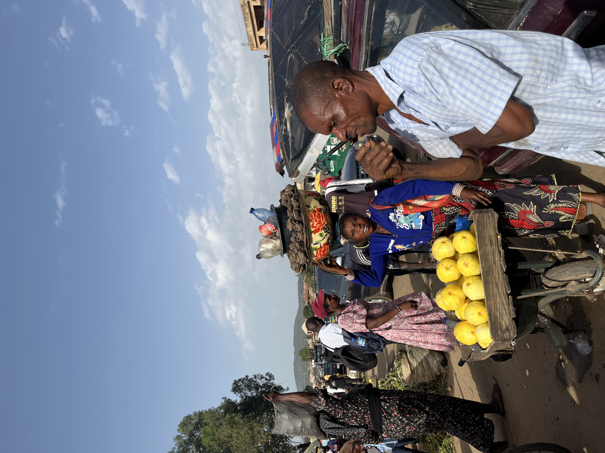 In Rafin Par, Nigeria, a busy outdoor market scene unfolds under a partly cloudy sky. In the foreground, a man in a light blue checkered shirt stands facing left, his arms crossed, holding what appears to be a small object to his mouth. To his left, a woman in a vibrant blue jacket and patterned skirt smiles towards the viewer, her hands near a wooden cart filled with yellow fruits. Behind her, a woman in a pink dress and red cap walks away, while another woman in a black floral dress carries a large black sack on her head. Several vehicles are parked or moving in the background, indicating the market is situated along a thoroughfare. Other market-goers and stalls are visible, suggesting a lively commercial environment. The ground is dirt, and trees and buildings are visible in the distance.