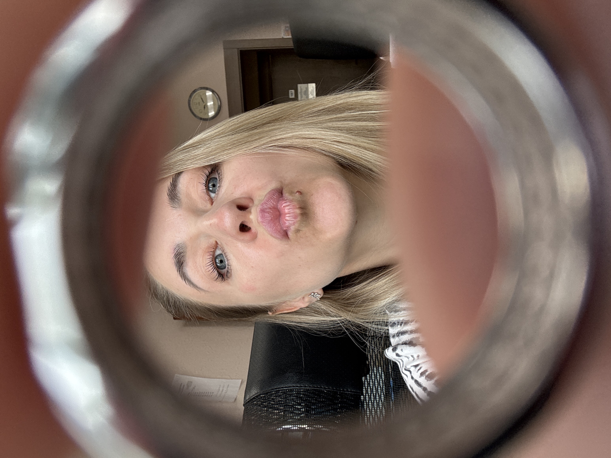A selfie photograph shows a blonde woman looking through a circular object, possibly a metal pipe or tube. Her face is close to the camera with pursed lips, fair skin, and blue eyes. She is wearing a small earring. Parts of her shirt can be seen. In the background, out of focus, there is a clock on the wall, a doorway, and a dark object that might be a chair. The image does not provide enough information to confirm that it was taken in Chaikovsky, Russia.