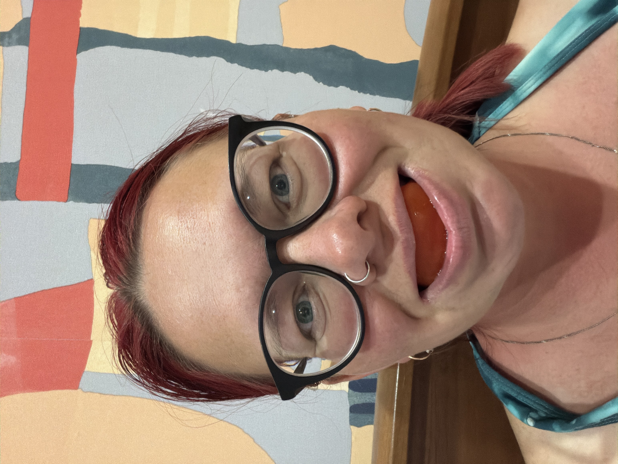 A woman with red hair and black glasses is holding a tomato in her mouth. She has a nose ring and earrings, and is wearing a teal top with a silver necklace. The background consists of a wall with a geometric pattern and a wooden frame. The location is likely indoors. The city of Chaikovsky, Russia is not discernable from the image.