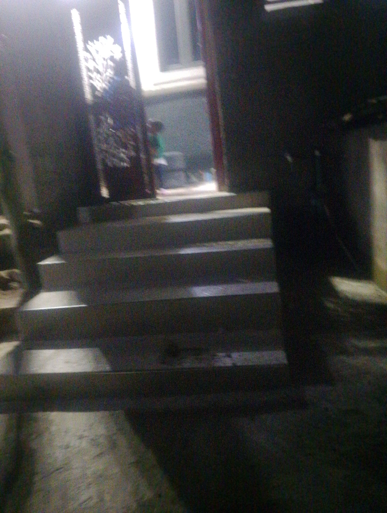 The image depicts a nighttime scene of an outdoor staircase leading to an entrance, likely a residential or commercial building. The staircase is made of concrete and appears to have at least five steps, with each step being wide and shallow. The entrance area above the stairs is dimly lit, with a bright light emanating from within the building, casting shadows across the steps and the surrounding area.
To the left of the stairs, there is a dark, textured wall, possibly made of stone or rough concrete. A portion of a person, seen from the waist up, is visible behind a decorative, ornate partition or gate structure. This person is wearing a green top. The partition itself is dark with intricate patterns and appears to be a prominent decorative element near the entrance.
The ground in front of the stairs is a rough, uneven surface, suggesting it is a pathway or courtyard. The overall impression is of a slightly run-down or older building, given the grainy quality of the image and the apparent wear on the surfaces. The lighting suggests it is nighttime, and the environment appears to be outdoors.
While the exact location in Jos, Nigeria, cannot be determined from the image alone, the presence of such an architectural style, particularly the decorative partition, can be common in residential areas across Nigeria. The scene is consistent with the exterior of a home or a small business in a city like Jos, characterized by its urban environment and likely a mix of traditional and modern architectural influences.