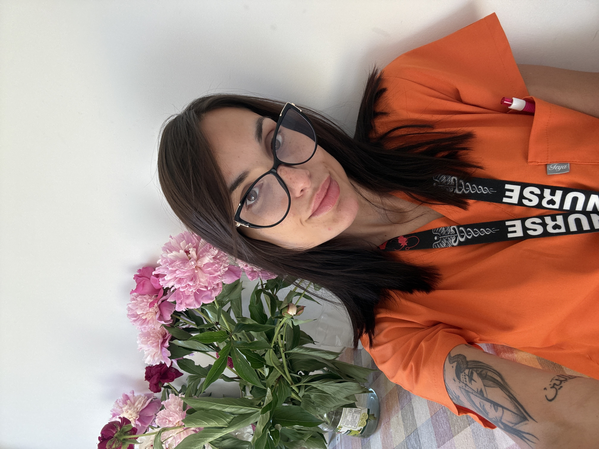 A woman with dark hair and glasses poses for a selfie in front of a bouquet of pink and burgundy peonies. She wears an orange scrub top with a "Nurse" lanyard around her neck and has tattoos on her left arm. The background is a white wall, and a glimpse of a patterned tablecloth is visible. The location is possibly Pleven, Bulgaria, as the woman has made posts indicating that this is her place of residence.