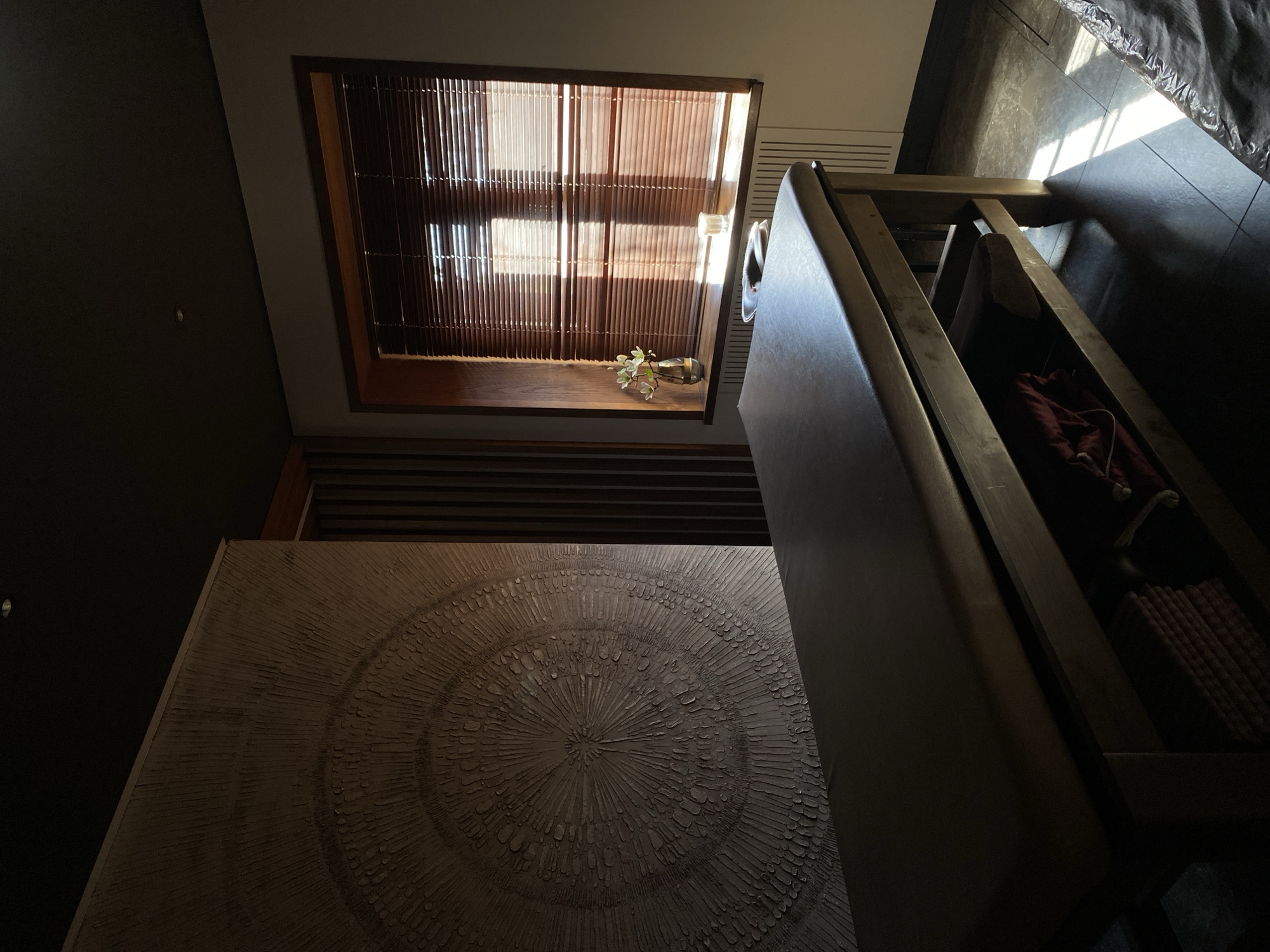 A high-angle shot captures a dim interior with several notable elements. A square window with bamboo blinds and a small vase containing foliage is set into a wall. A textured, circular design is visible on a nearby surface. A dark brown, cabinet-like structure dominates the right side of the frame. The floor is tiled with dark gray tiles. The environment appears dimly lit. It is not possible to determine the location of the photograph as Votkinsk, Russia. The presence of people is not visible.