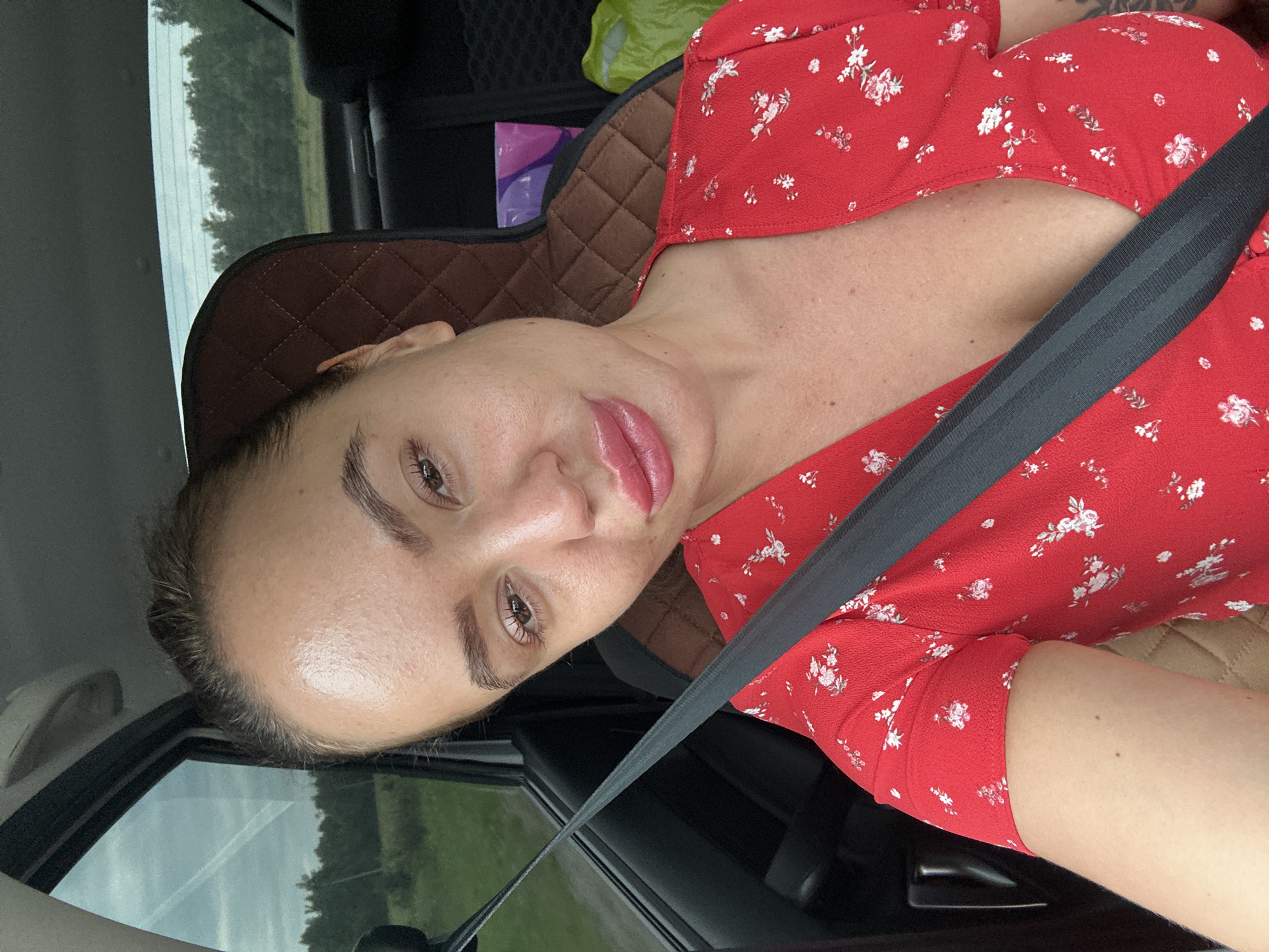 A woman, likely in her late twenties or early thirties, is seen in a car, possibly on a road near Uvel'skiy, Russia. She is wearing a red dress with a floral pattern and a black seatbelt across her chest. Her hair is pulled back, and she is looking directly at the camera with her lips slightly parted. The interior of the car is visible, including a quilted seatback and a window showing trees and a cloudy sky. There are bags in the back seat. The overall lighting suggests it is daytime.