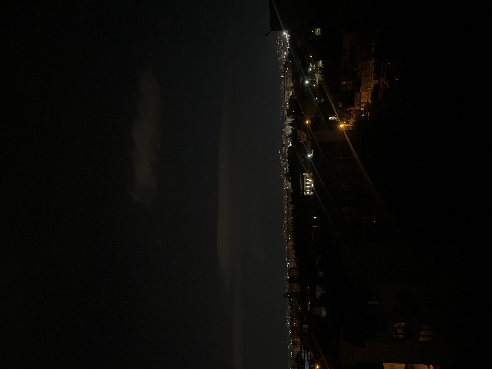 The image displays a wide, horizontal panoramic view of a city at night, appearing rotated 90 degrees clockwise in its original orientation. The scene captures a vast urban landscape under a dark sky, observed from an elevated vantage point, likely in Sofia, Bulgaria.
The upper half of the image (left side in the rotated original) consists of the deep black of the night sky. Faint, wispy cloud formations are subtly visible within this expanse, softly illuminated, and a few tiny, indistinct specks of light, possibly stars, dot the darker areas.
A distinct, glowing horizontal line marks the horizon, separating the dark sky from the brightly lit city below. The lower half of the image (right side in the rotated original) showcases a sprawling urban area, defined by a dense cluster of artificial lights. These lights, primarily warm yellow-orange from streetlights and cooler white from buildings, extend across the entire frame, signifying a large settlement stretching into the distance. Individual buildings are largely obscured by darkness and distance, appearing as silhouettes highlighted by their own illumination. Along the extreme right edge, a dark, indistinct structure or railing is partially visible, indicating the photograph was taken from a high balcony or rooftop.
No people or specific activities are visible in this static night shot. The time of day is clearly night, and there are no discernible adverse weather conditions. No visible text is present in the image.