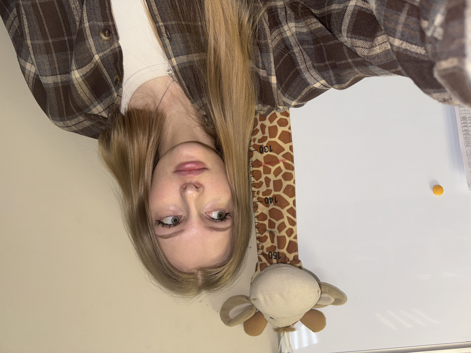 A selfie-style, slightly tilted photograph depicts a young woman standing next to a wall-mounted giraffe height chart. The woman is positioned on the left side of the frame; her head is at the top of the image, and her torso is partially visible. She has light blonde hair, fair skin, and her eyes are glancing to the side. She is wearing a brown and tan plaid shirt over a light-colored top.
To her right is a height chart in the form of a giraffe, with the numbers "130", "140", and "150" visible. The bottom part of the giraffe is a stuffed plush toy.
The background appears to be a plain white wall with a yellow object in the upper right corner. There is a vertically oriented piece of paper affixed to the wall to the right of the frame.
The location is the city of Perm, Russia.